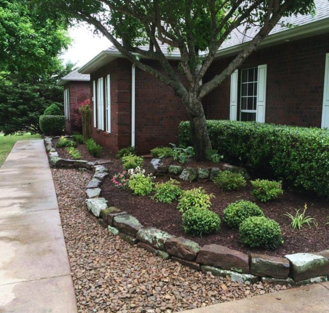 A well-maintained garden bed with shrubs and small plants, bordered by large rocks, along a concrete sidewalk outside a brick house with white window shutters and a large tree providing shade.