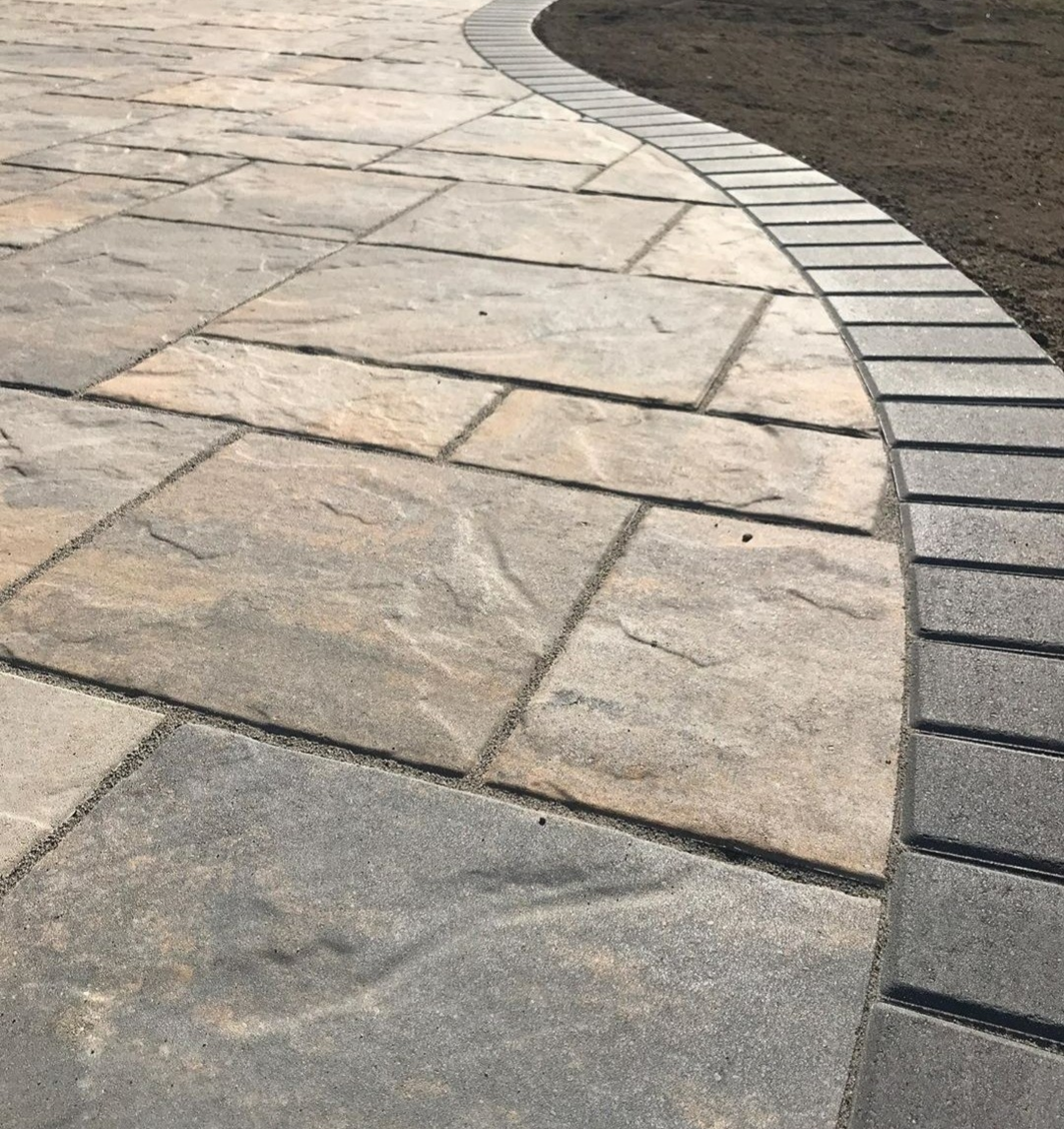 Curved paver patio featuring natural stone slabs with a contrasting brick soldier-course border for clean edges and long-term durability. This custom hardscape design enhances drainage, prevents shifting, and creates a smooth transition between patio