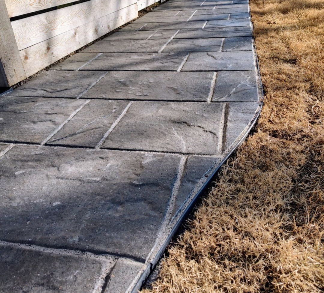 Custom stone-look concrete walkway featuring large rectangular slabs with a natural slate texture and clean edging for long-term durability. This hardscape design creates a smooth, functional transition along the home’s exterior while controlling ero