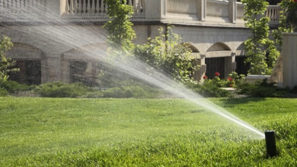 A sprinkler watering a well-maintained green lawn in front of a large building with balconies and plants.