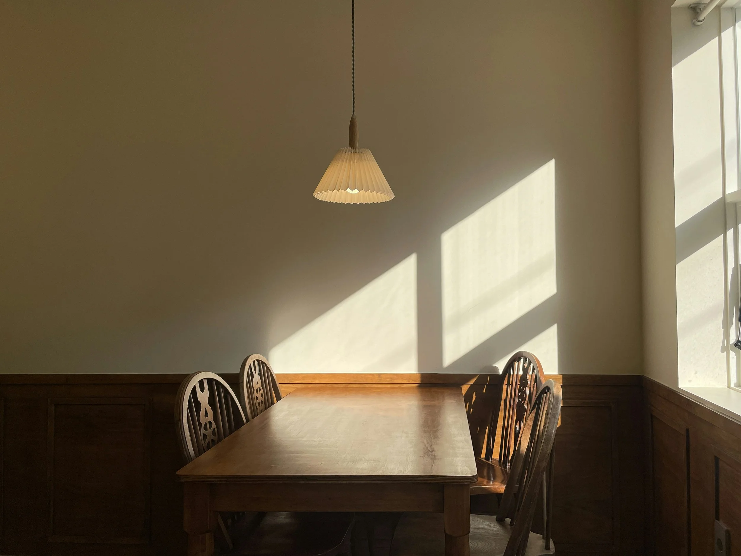 A wooden dining table with six chairs around it, shadowed in a room with sunlight streaming through a window, creating diagonal shadow lines on the wall.