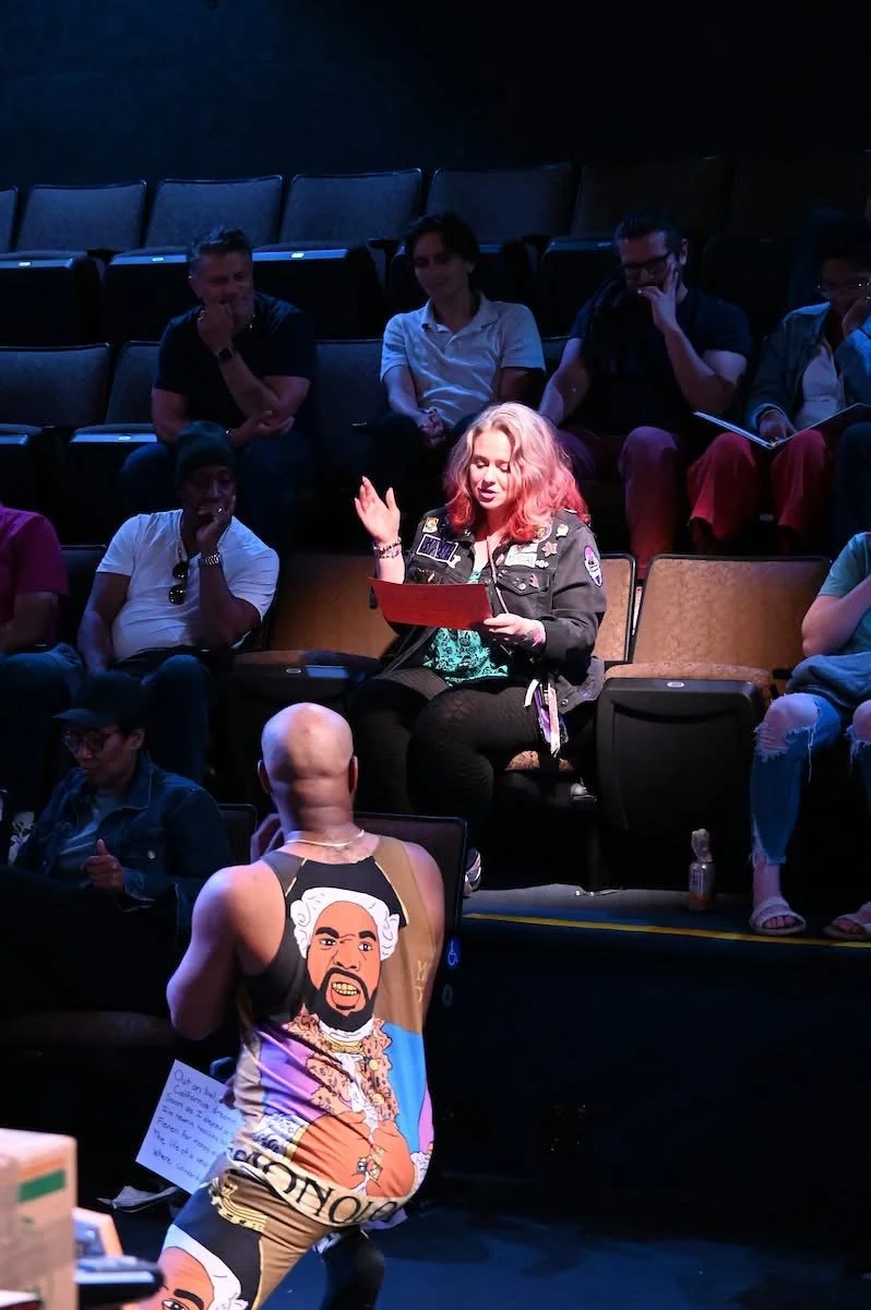 A group of diverse people sitting in a theater or auditorium, with a woman with pink hair in the center reading from a red paper, and a person with a bald head and cartoon character print on their shirt kneeling in front of her.