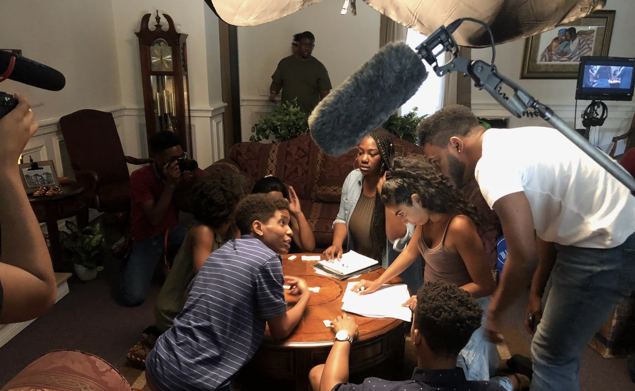 Group of people gathered around a table in a living room, engaging in a discussion or activity. A woman is pointing at papers on the table. Cameras and a boom microphone are set up, indicating a filming or recording session. The room features a grandfather clock, framed artwork, and various furniture.
