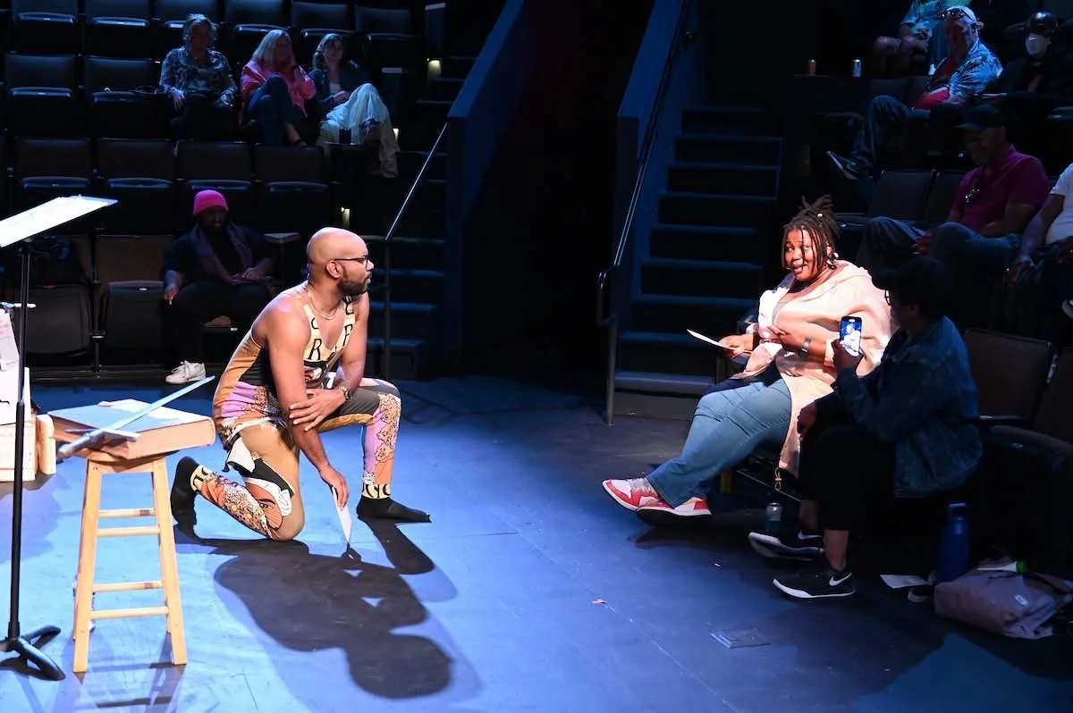 A man kneeling on stage with a paper in hand, speaking to two women sitting in the audience, one with a smartphone. Several people sit in dimly lit auditorium seats behind them.