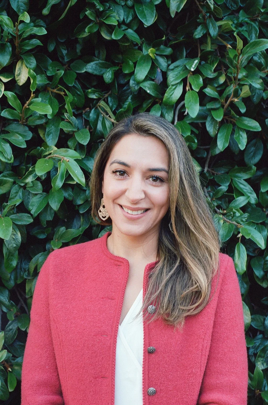 Gabriela is standing in front of a leafy background wearing a red  jacket.