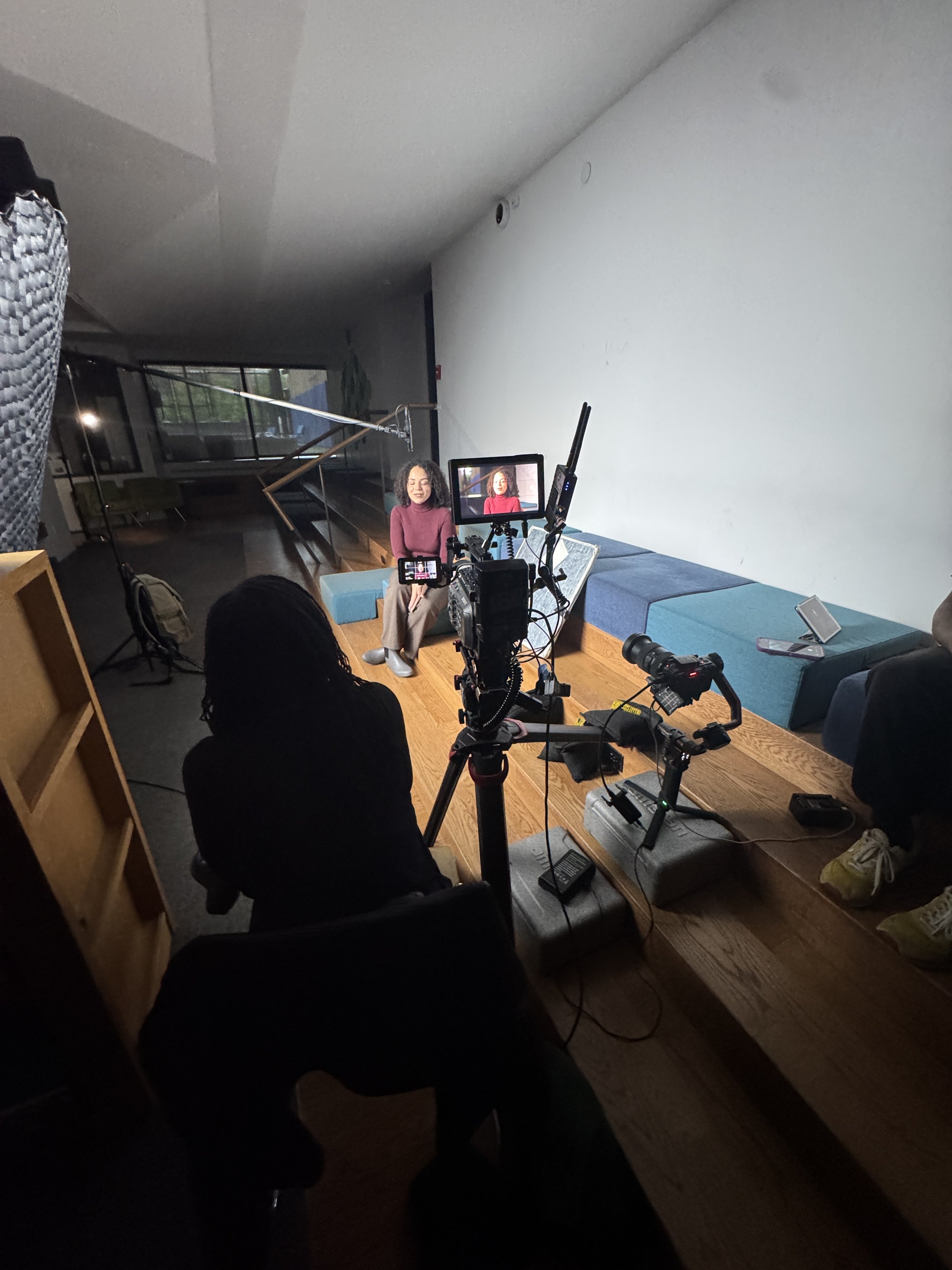 Film crew recording a seated woman in a studio living-room set with multiple cameras, monitors, and lighting equipment.