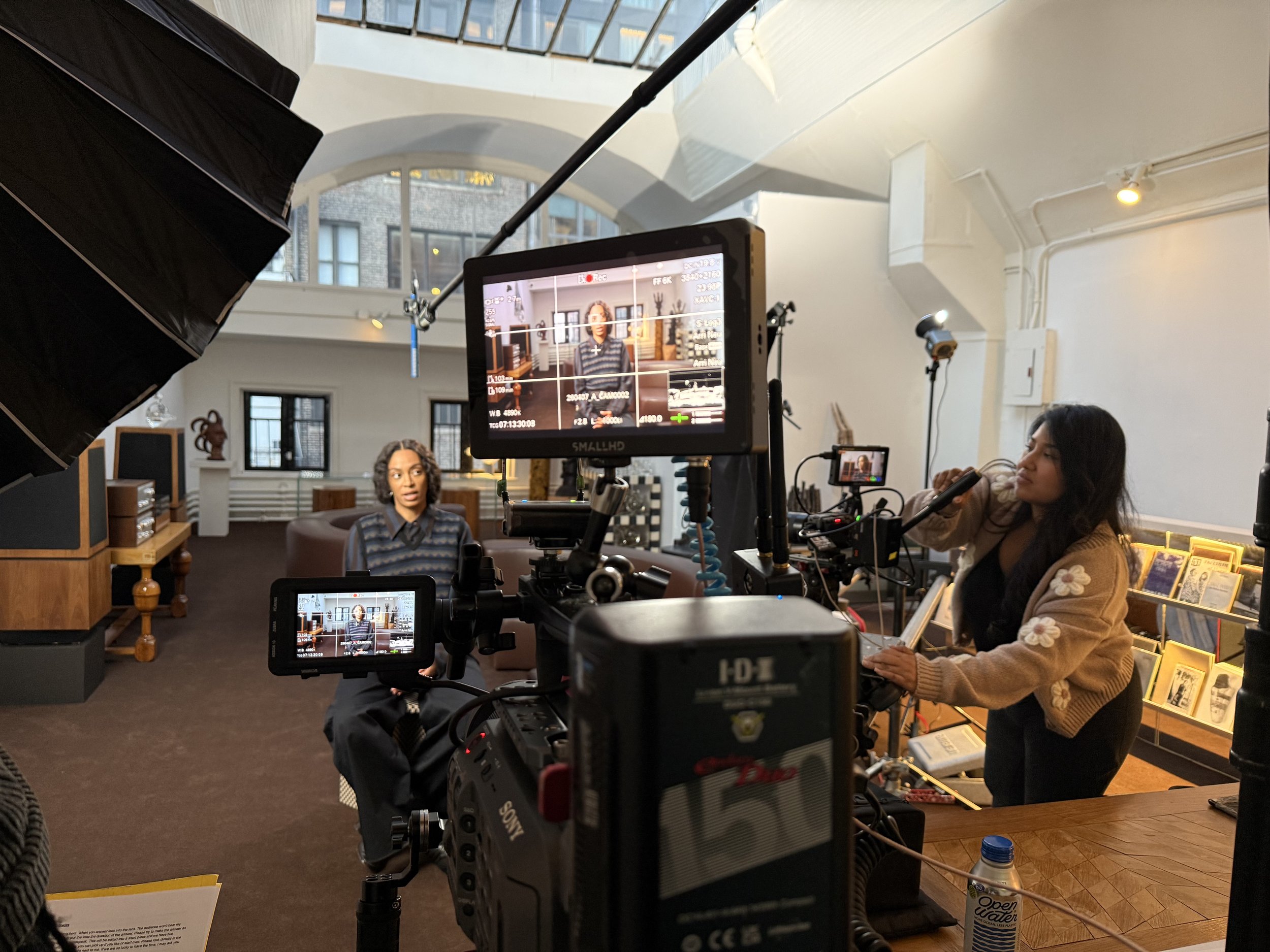 Behind-the-scenes film set with cameras, monitors, lighting equipment, a seated performer, and a crew member in a loft interior
