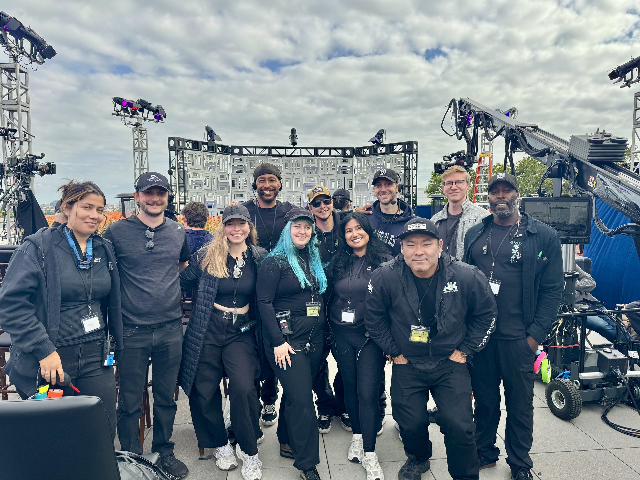 Film and television production crew posing for a group photo on an outdoor set surrounded by cameras, lighting rigs, and stage equipment.
