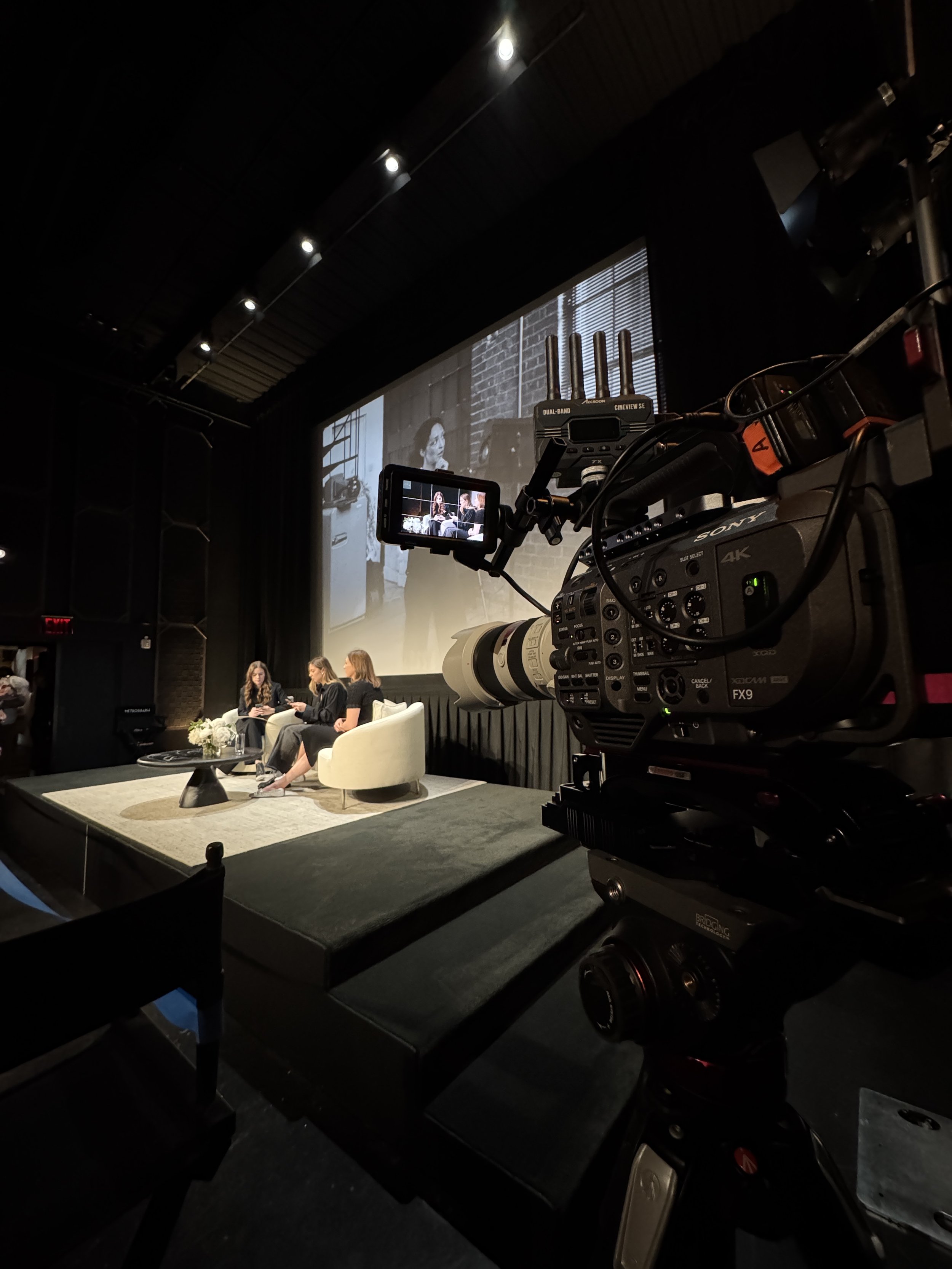 Professional Sony cinema camera filming three women seated on a stage beneath a projected screen in a dim studio.