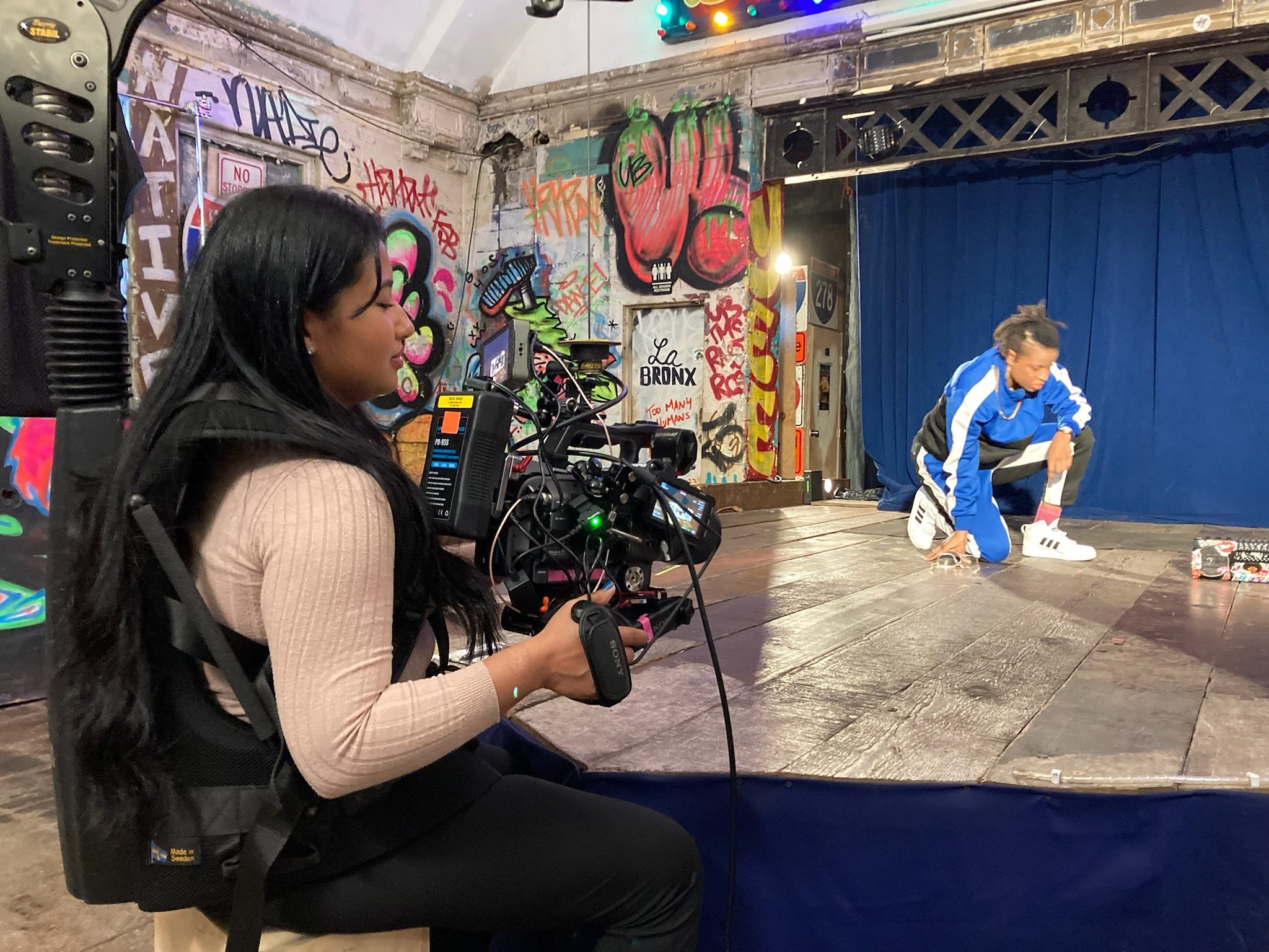 Videographer filming a dancer in a blue tracksuit performing on a graffiti-covered stage.