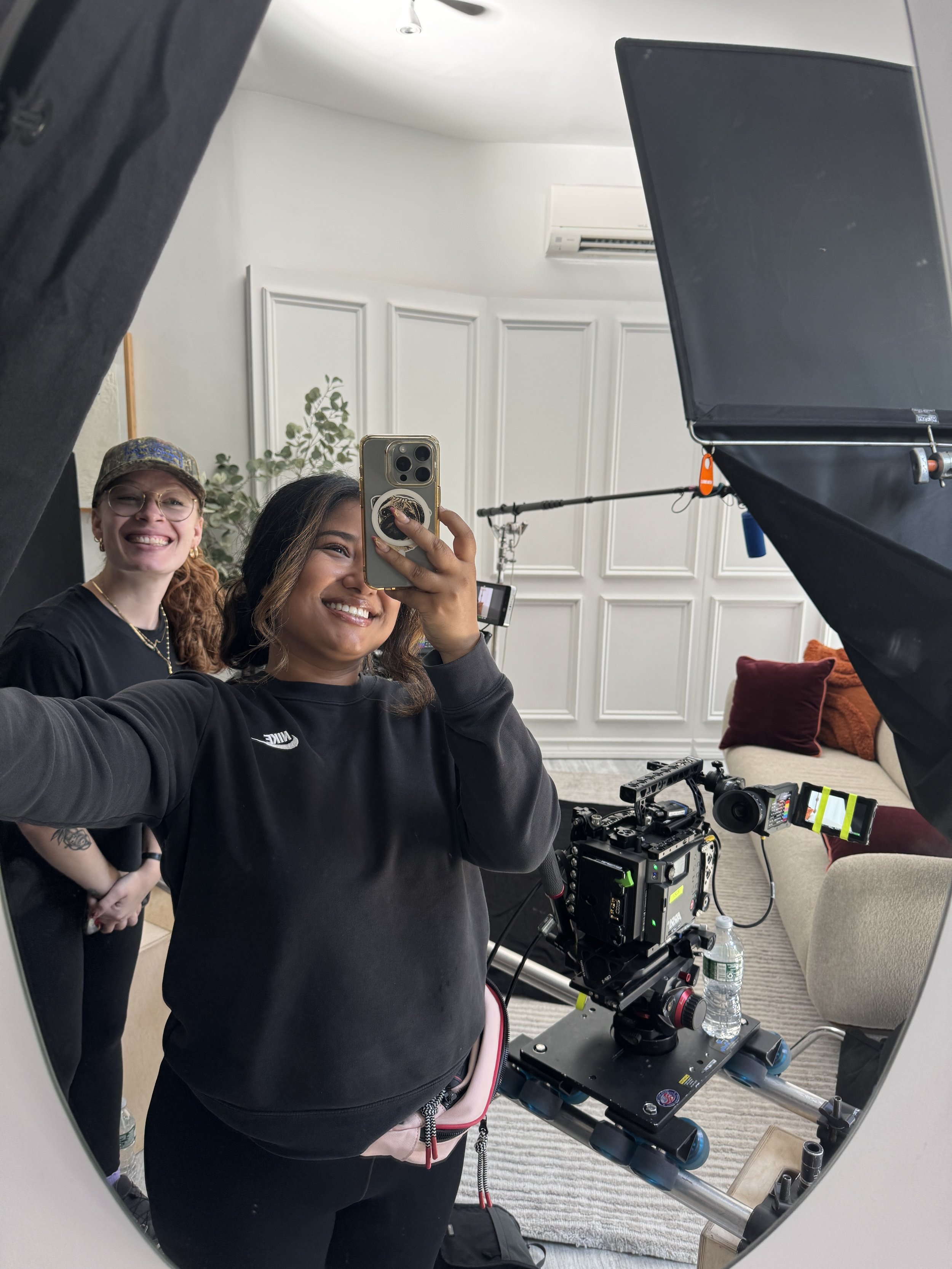 Two smiling women take a mirror selfie beside a professional cinema camera and studio lighting equipment.