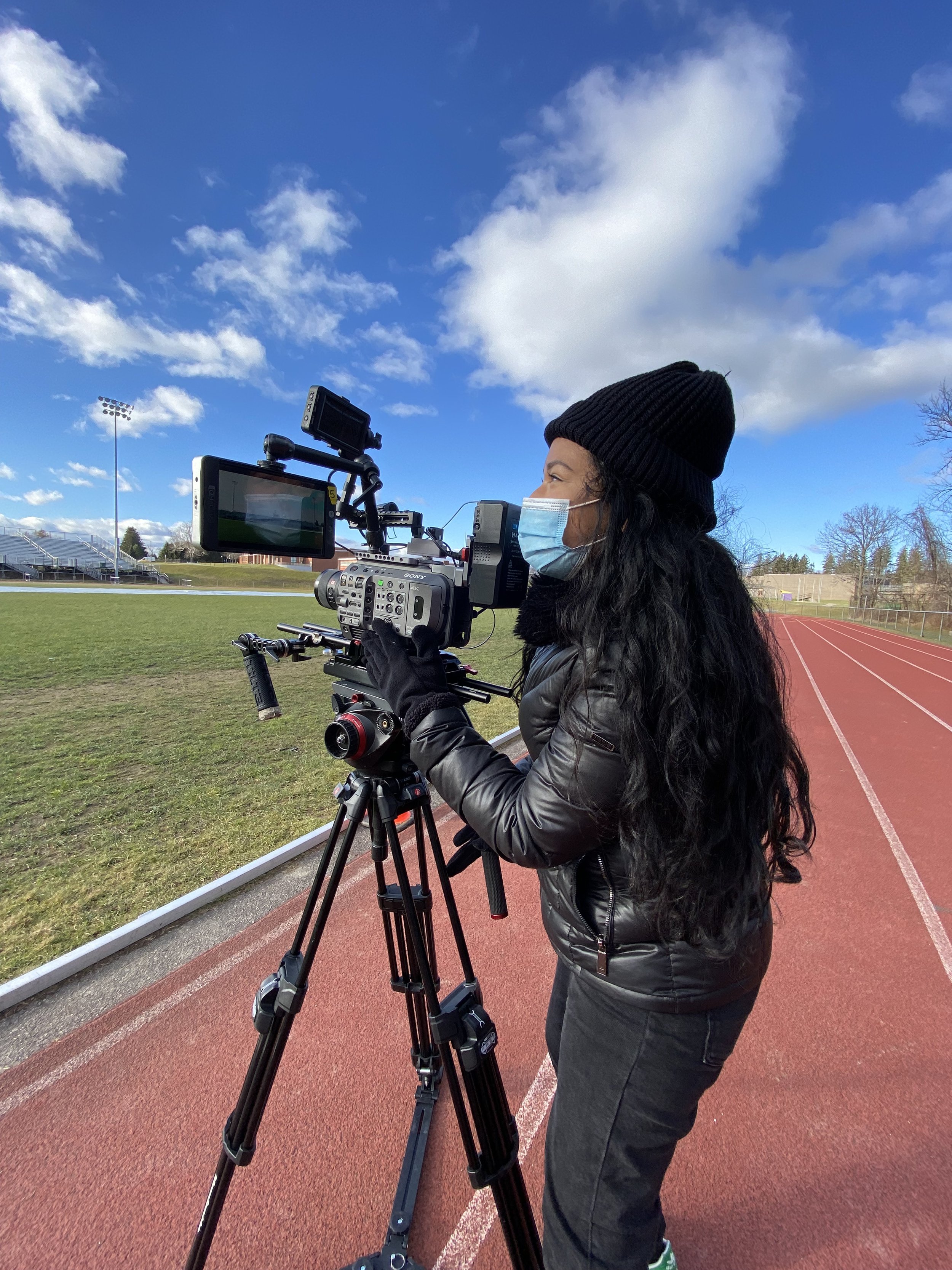 Masked videographer operating a professional camera on a tripod beside a running track and athletic field.
