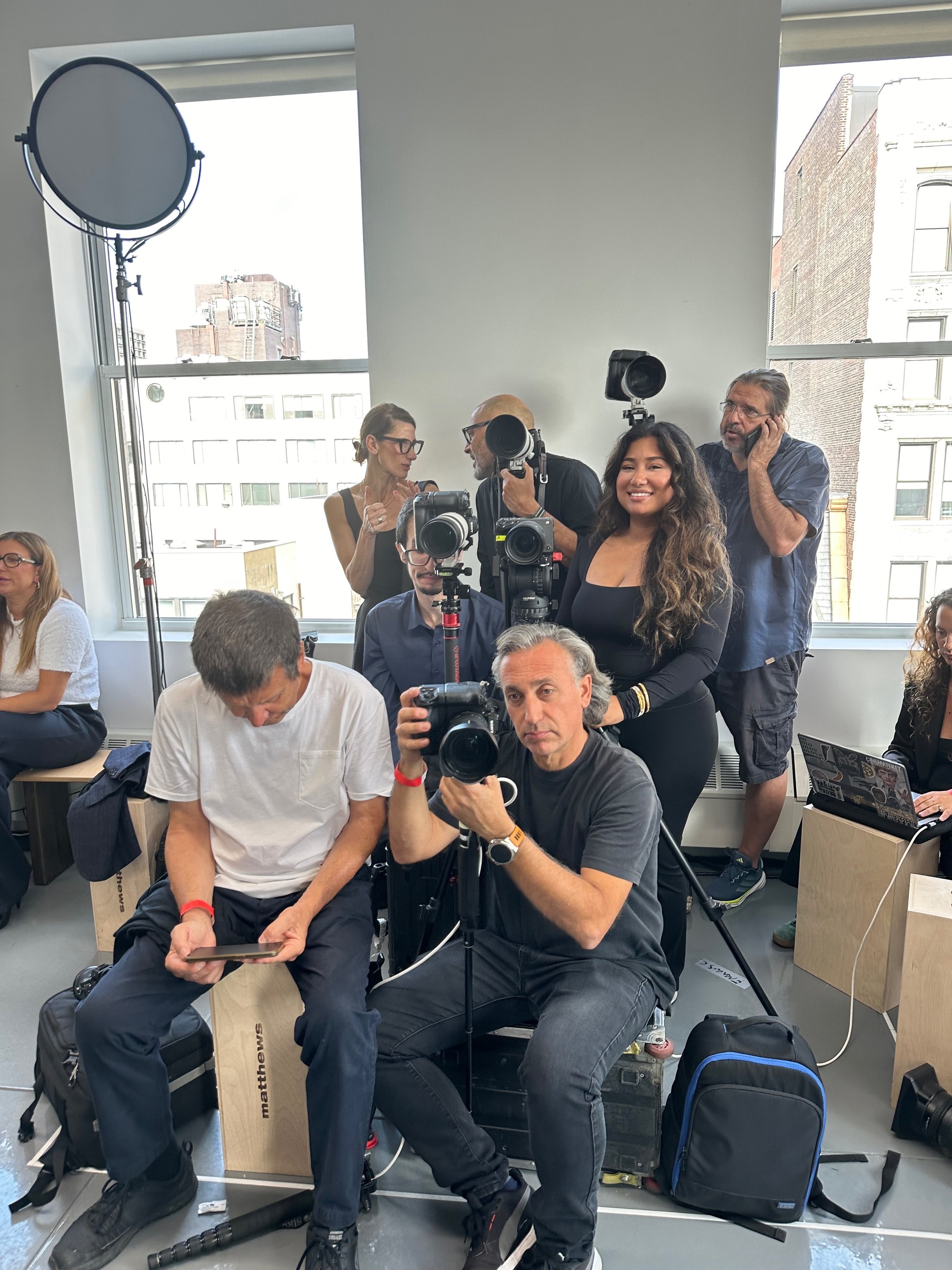 Film crew with cameras and lighting equipment gathered in a bright studio overlooking city buildings