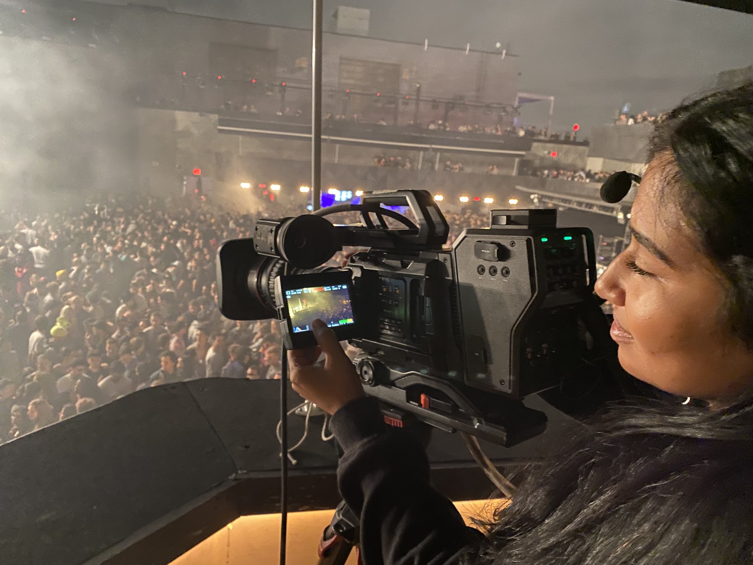 Camera operator filming a crowded indoor concert venue from a balcony.