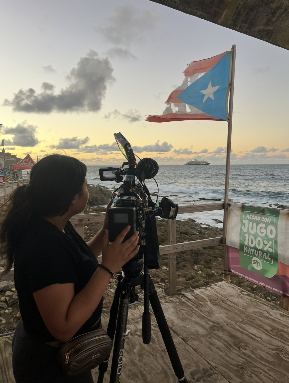 Videographer filming the ocean at sunset beside a Puerto Rican flag and natural juice sign