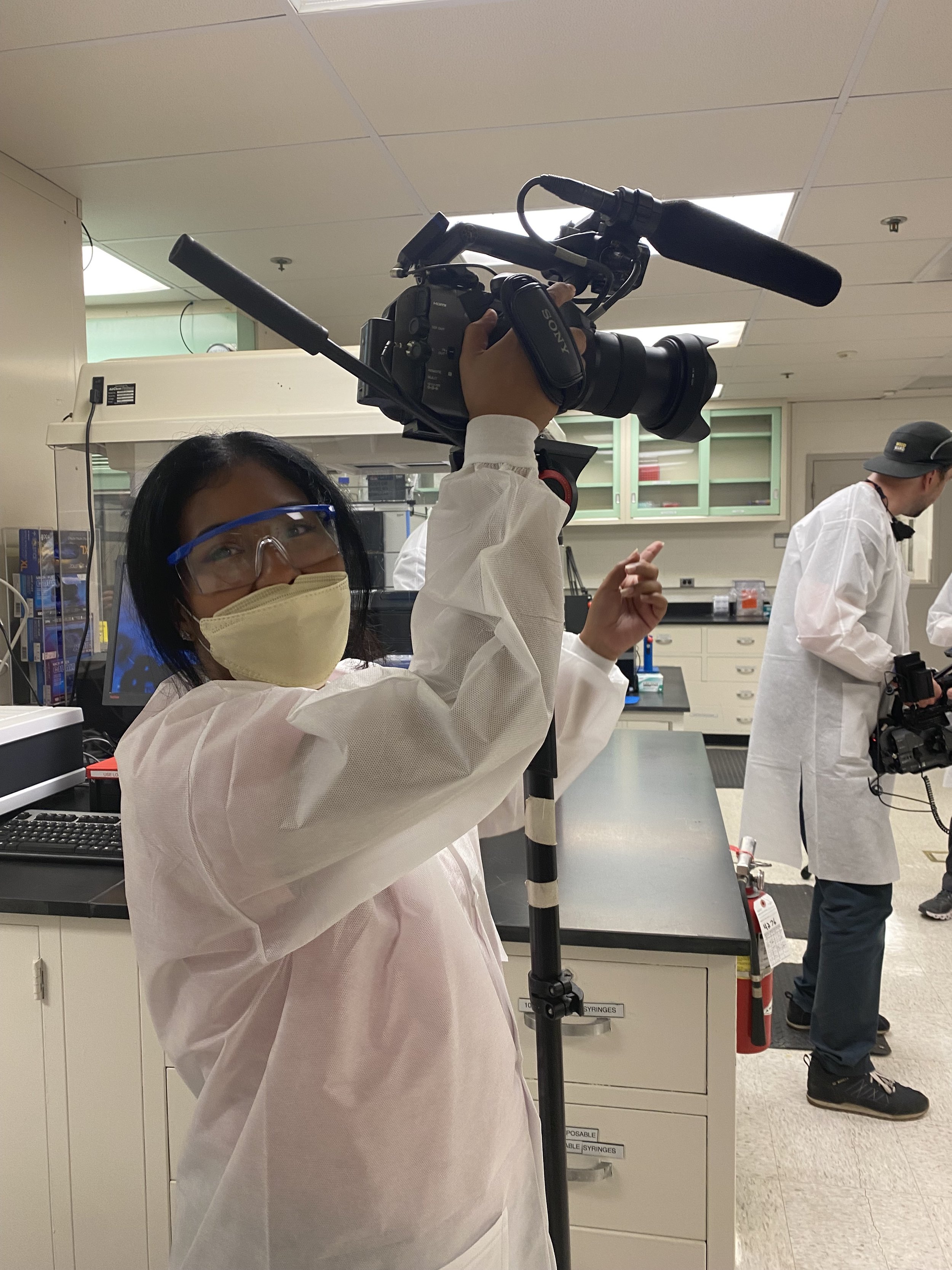 Film crew in protective gowns filming inside a laboratory with a professional Sony video camera.