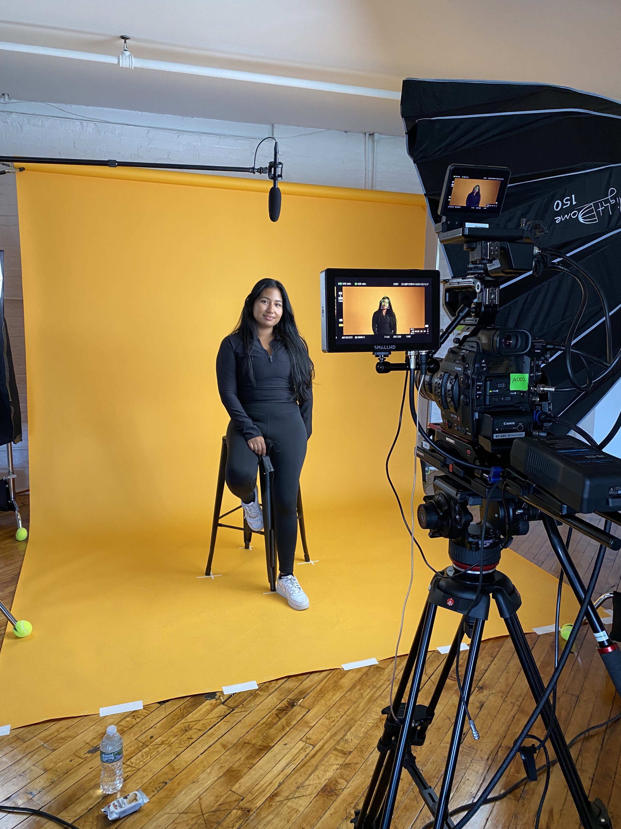 Behind-the-scenes studio shoot with a woman posing in front of a yellow backdrop, professional camera, monitors, lighting equipment, and boom microphone.