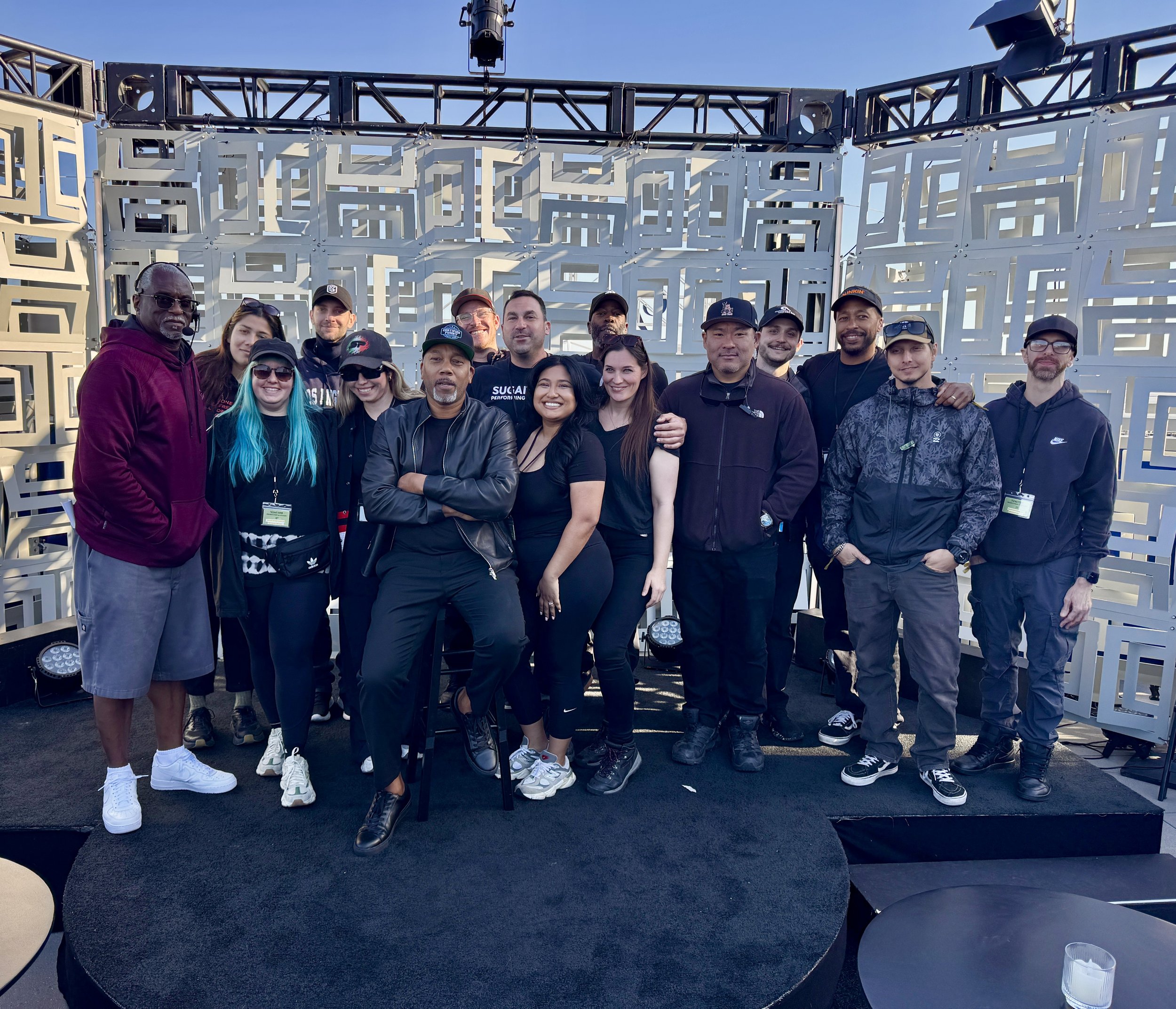 Group portrait of event staff and performers on a stage in front of a geometric white backdrop