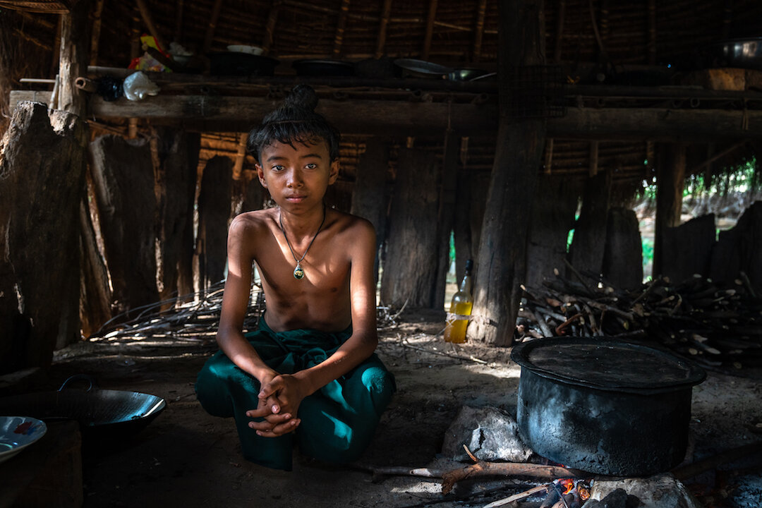 A young boy with the traditional hair knot sitting in a twig hut, the "hidden" village