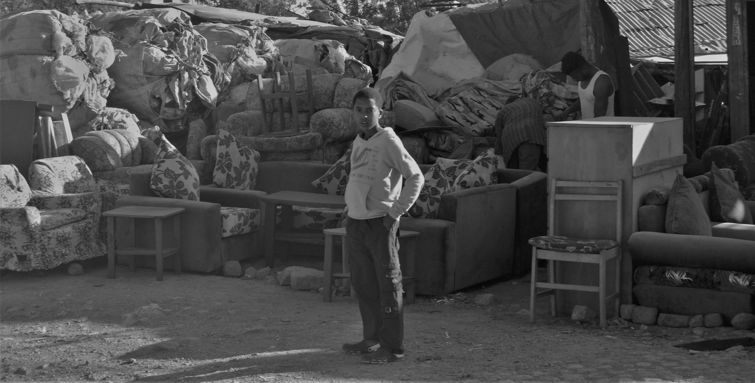  11. In this photo, a young boy stands proud in front of his family's used furniture shop. He stares into the camera with a smile, proud of the hard work he has put in to ensure his family's business is successful. The store has several pieces of fur