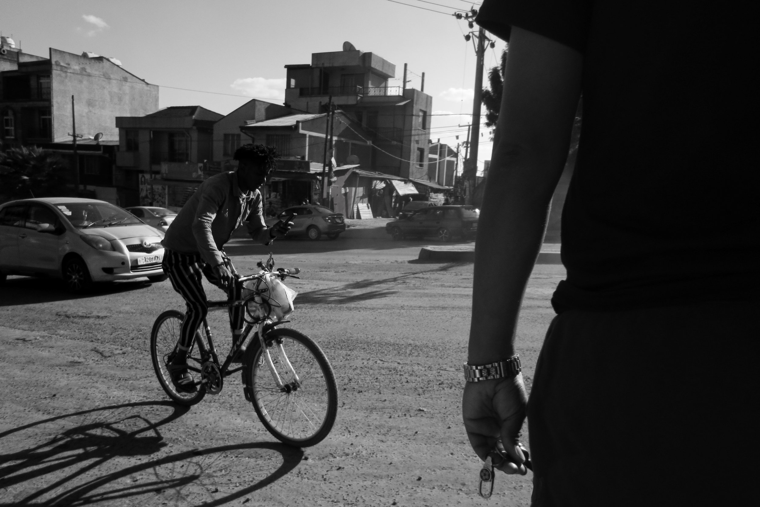  18. A young man is riding his bicycle along a road, engrossed in checking his phone with one hand as he pedals with the other. With the sun setting in the background. 