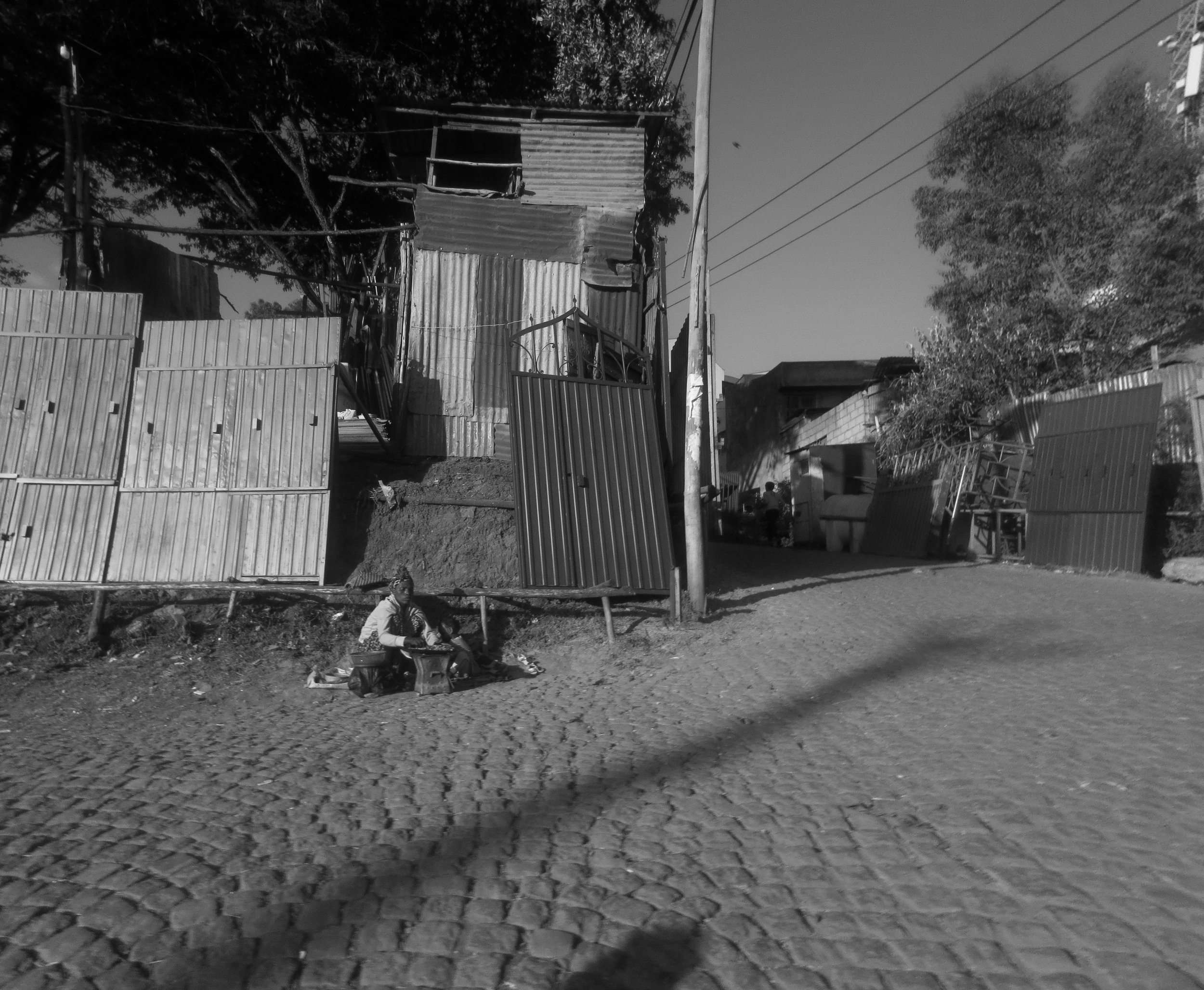  19. In the photo, a mother and her daughter are seen selling clothes by the side of a road, displaying various items of clothing such as shirts, dresses, and pants. The daughter, who looks to be around 10 years old, is sitting next to her mother, he