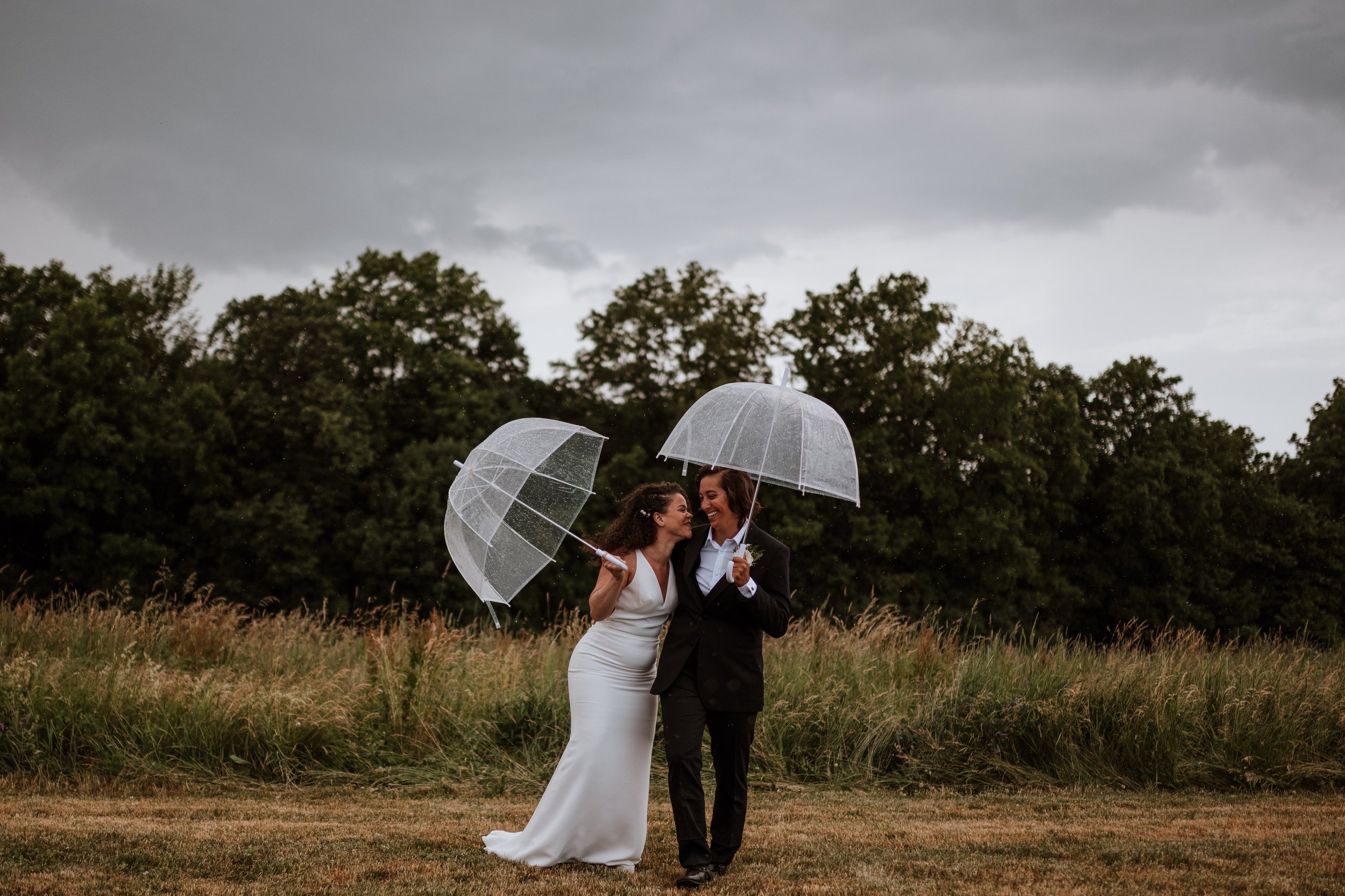 Newlywed lesbian couple laughing together under umbrellas while it's raining.