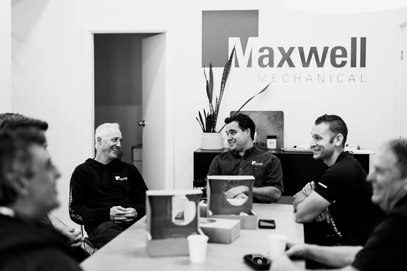 Black and white photo of the Maxwell Mechanical team sitting around a meeting table in their Kamloops office, with the company logo visible on the wall behind them.