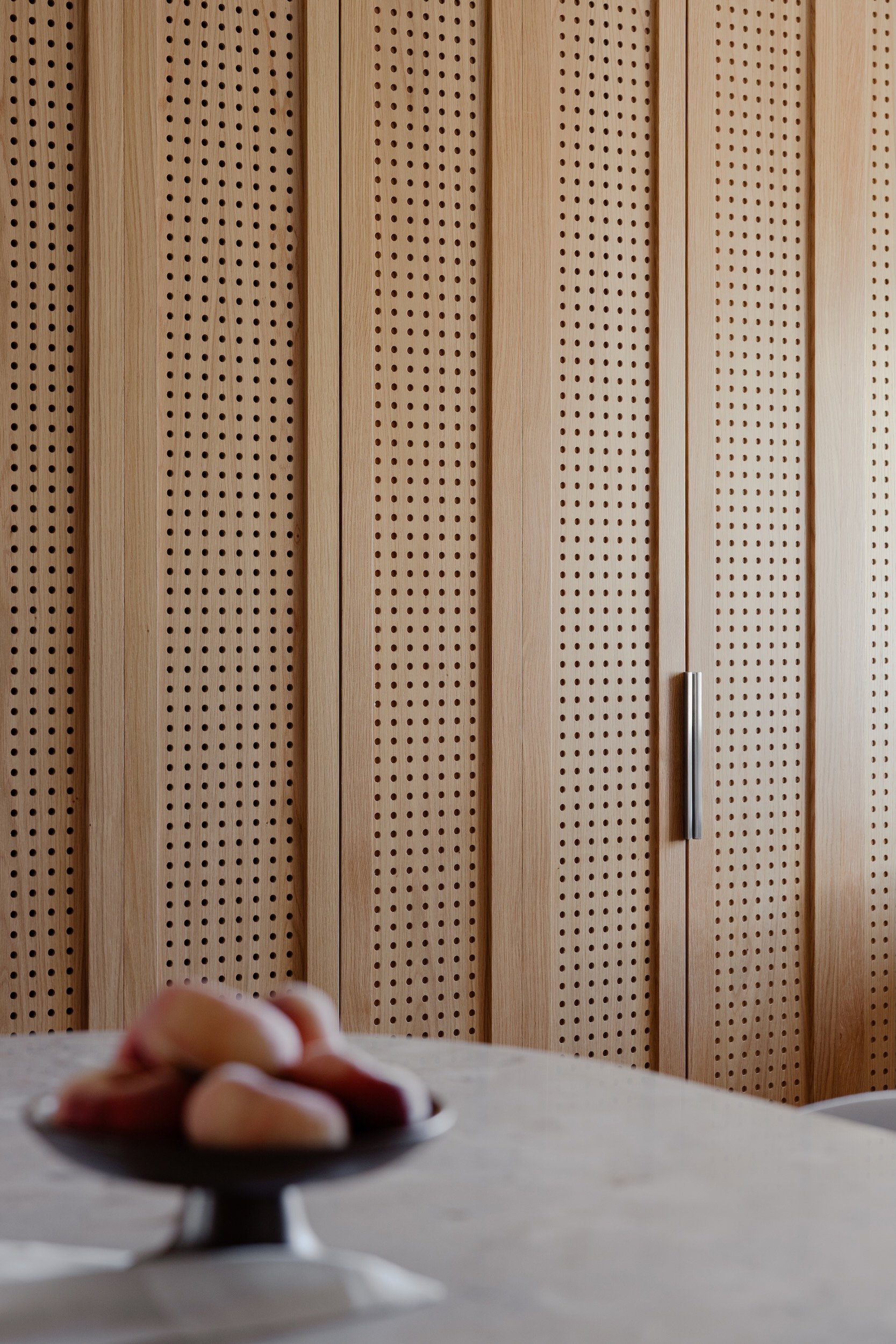 Lola Cwikowski Studio Cascais Residence project with close-up of custom perforated oak laundry cabinet doors; curved fluted oak island with Lioz countertop; footed fruit bowl with peaches.