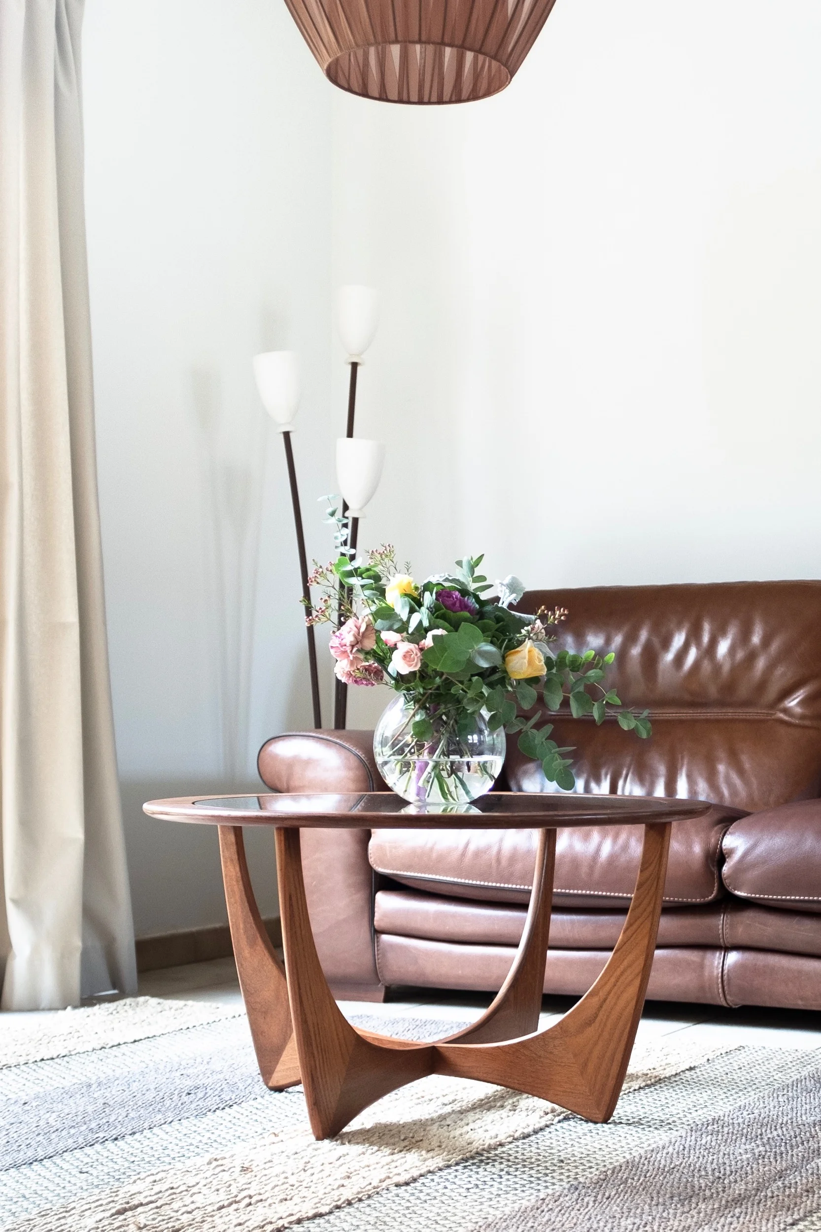Round vintage wood coffee table with flowers in front of leather couch and tulip lamp