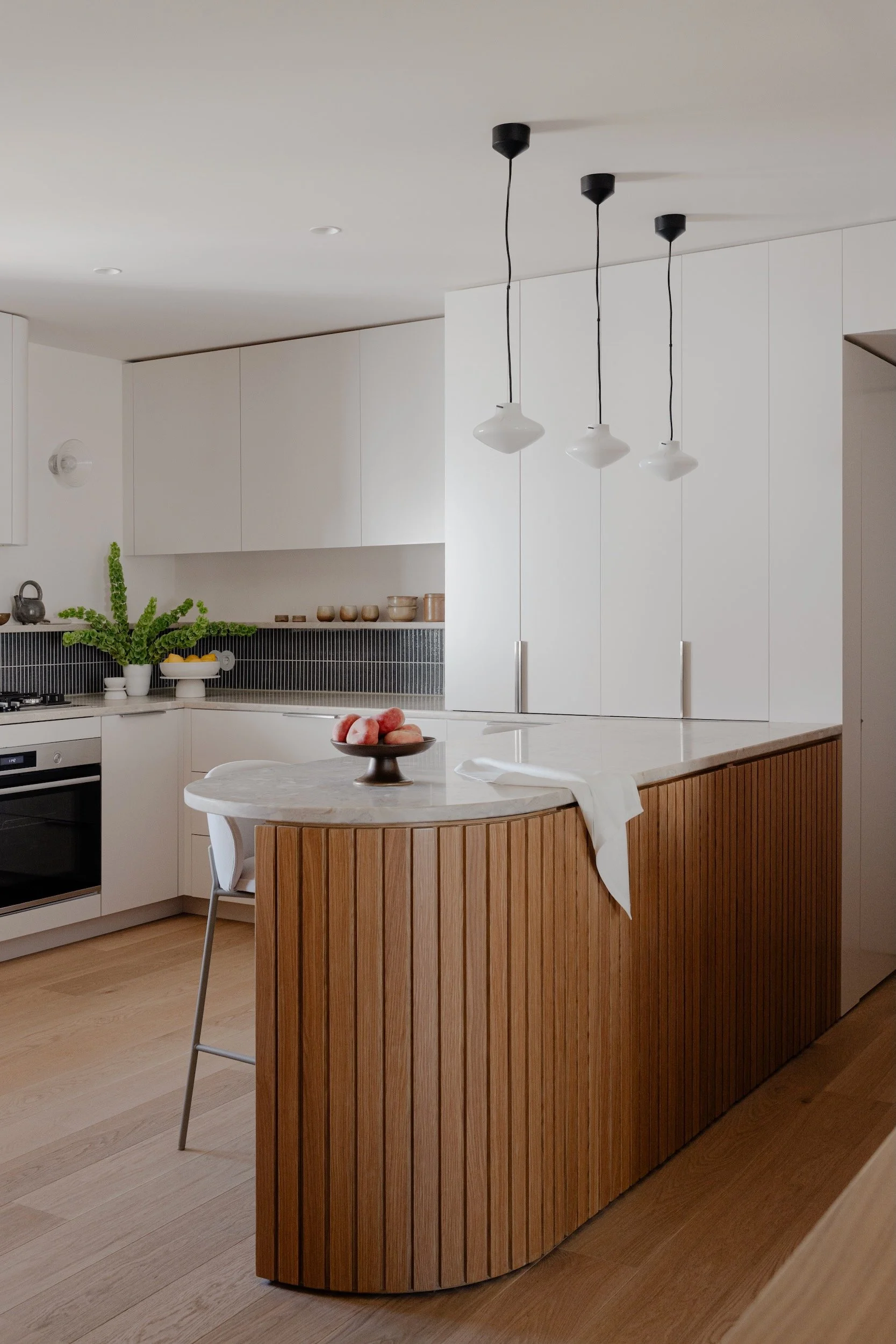 Lola Cwikowski Studio Cascais Residence project with custom curved fluted oak island with Lioz countertop; Lyfa Repose pendants; BoConcept Princeton stool; FDB Møbler U12 Sletterhage wall lamp; Marokk Kayo Tokyo tile backsplash; Bosch oven.