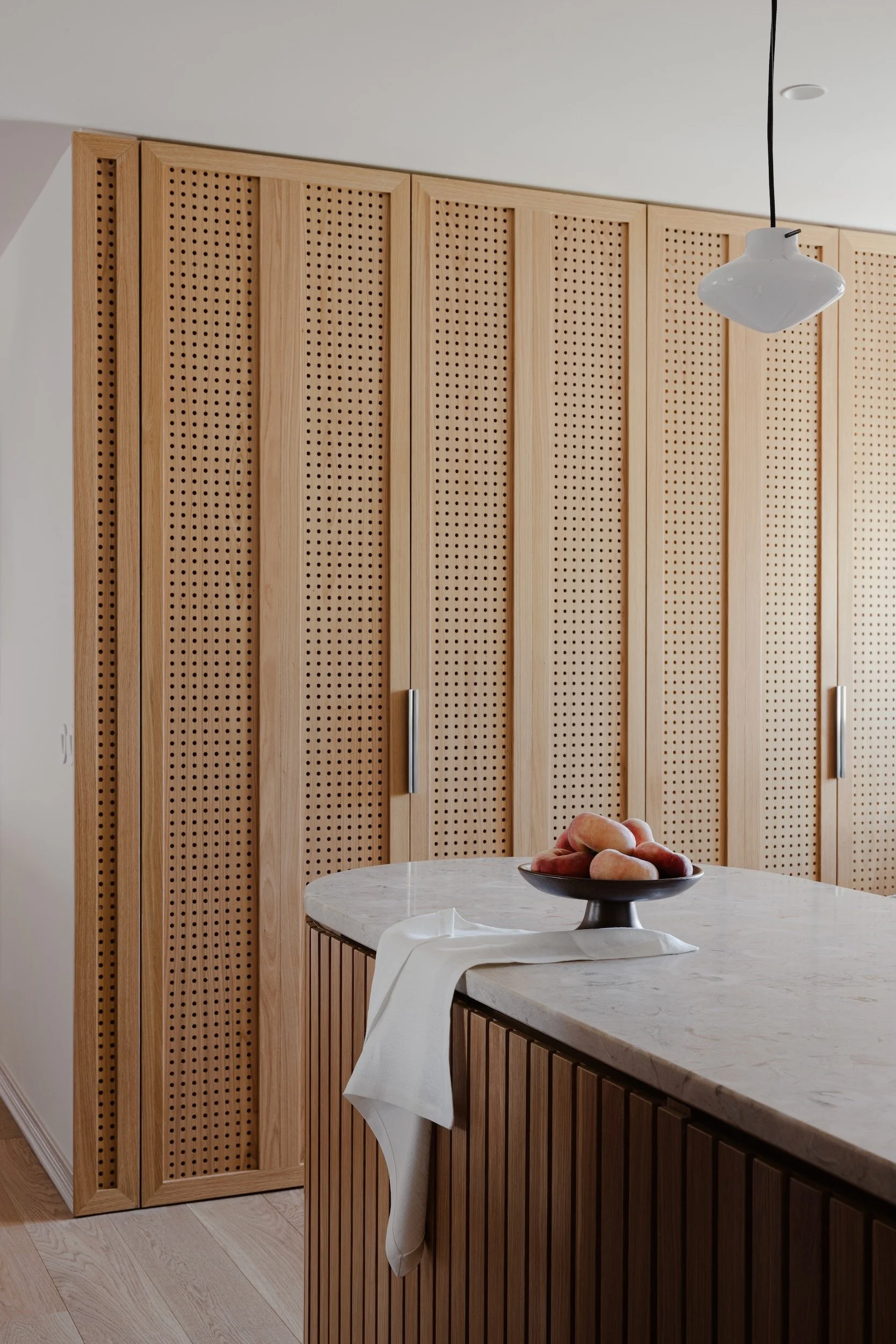 Lola Cwikowski Studio Cascais Residence project with custom perforated oak laundry cabinet doors; curved fluted oak island with Lioz countertop; Lyfa Repose pendant; footed fruit bowl; linen cloth.