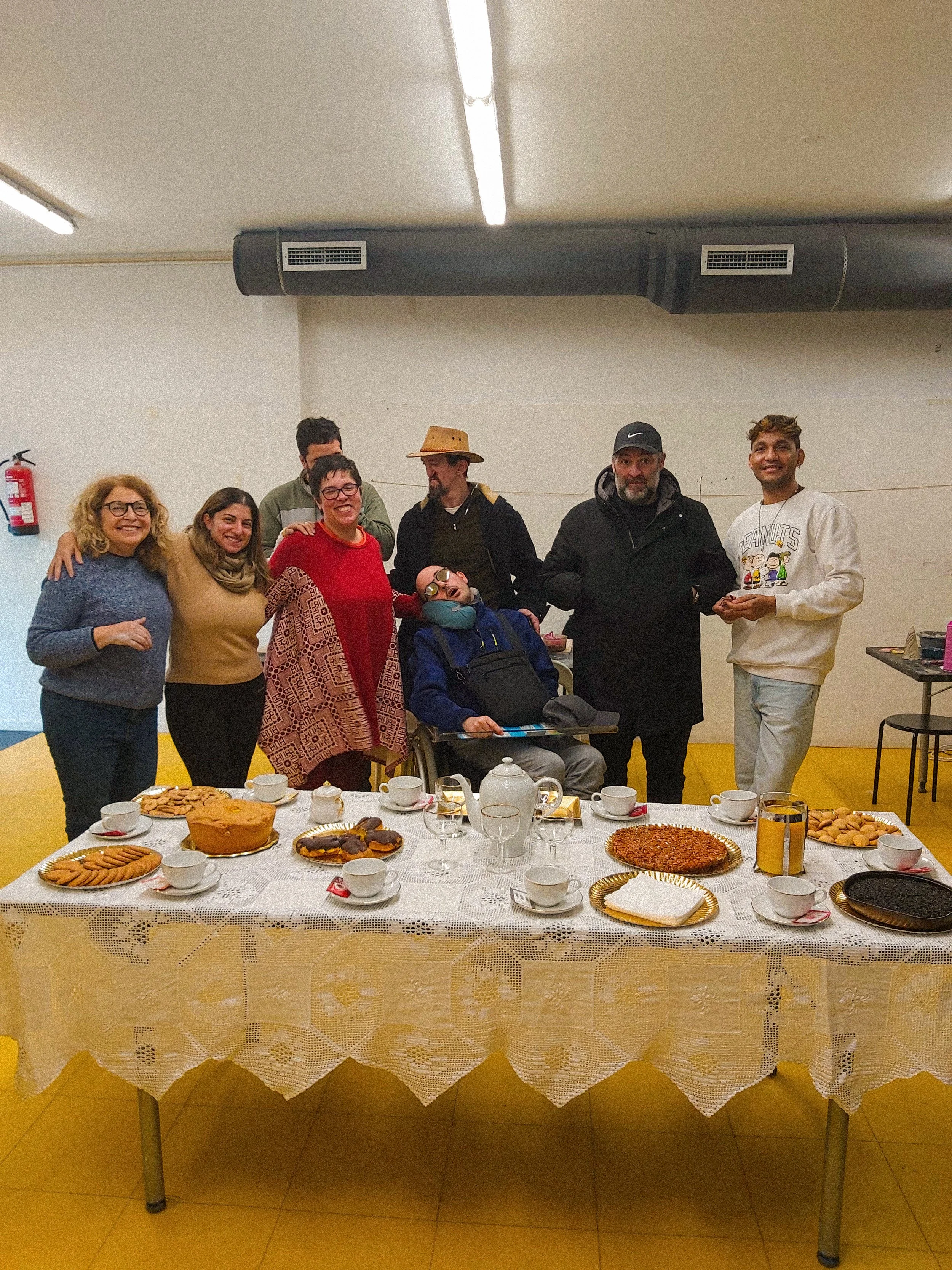 Grupo de pessoas ao redor de uma mesa com alimentos, celebrando um momento juntos, incluindo uma pessoa em cadeira de rodas, todos sorrindo e posando para a foto.