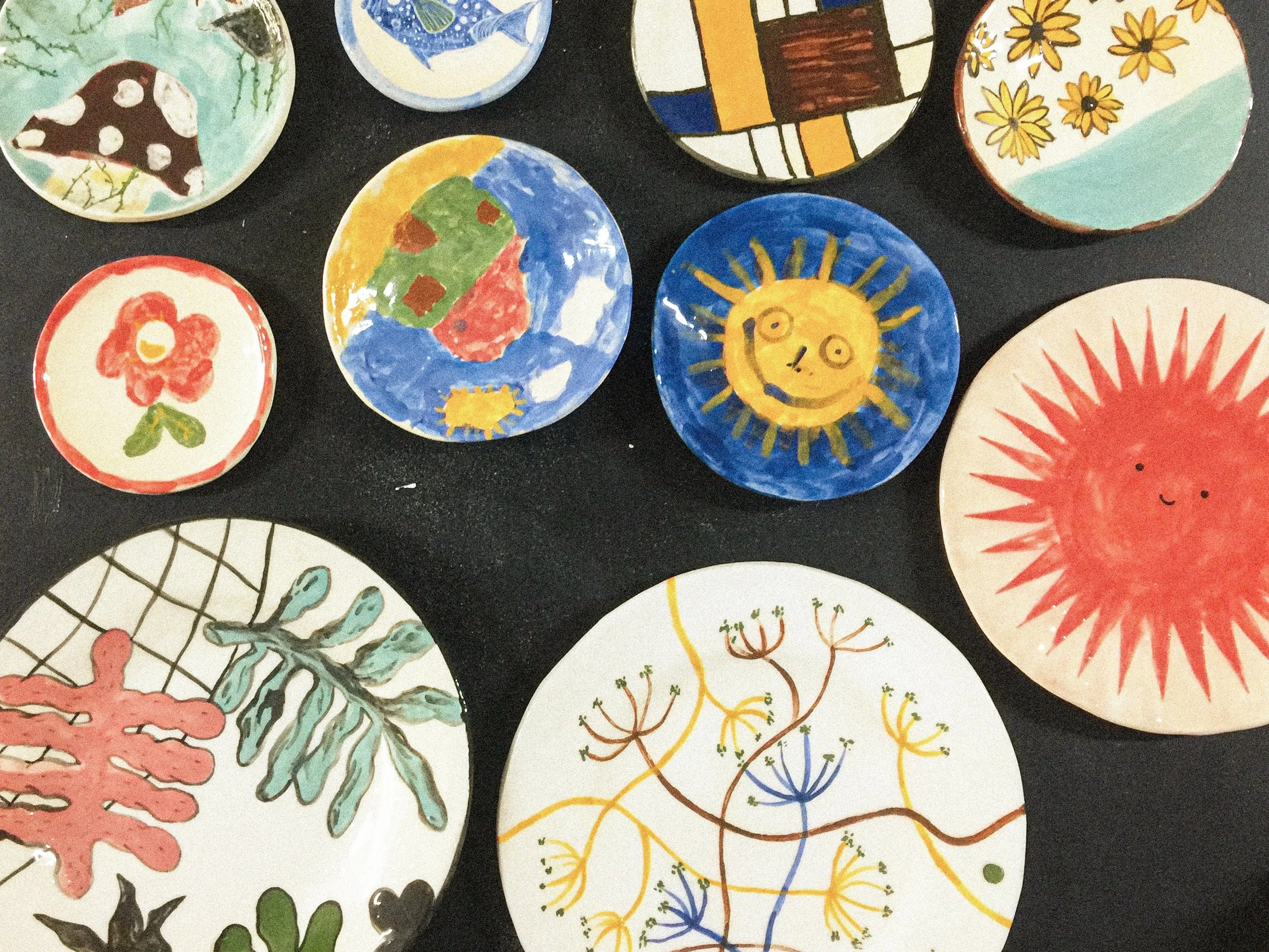 Vários pratos de cerâmica pintados com diferentes desenhos coloridos, incluindo o sol sorridente, a Terra, flores, folhas, animais e padrões abstratos.