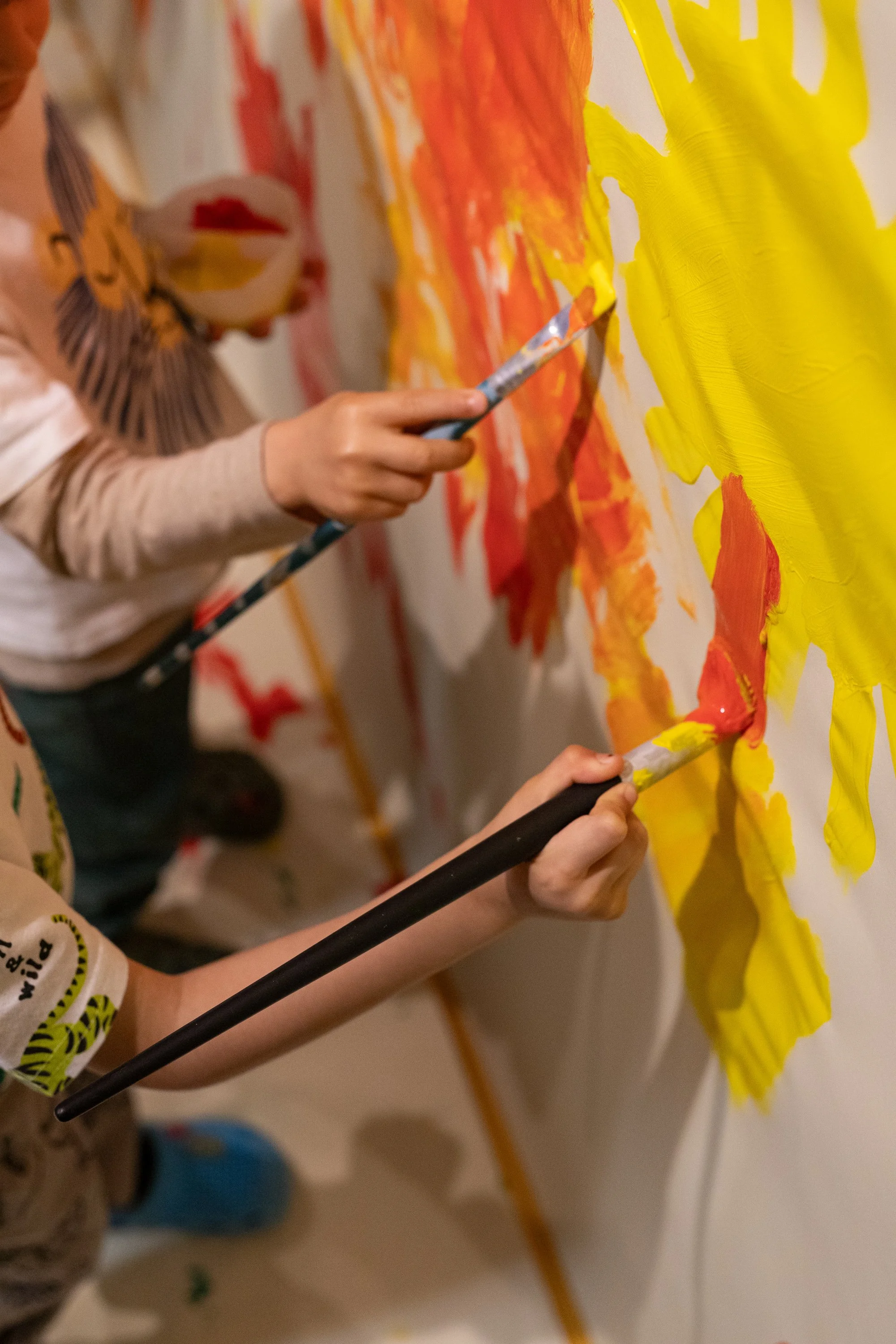 Crianças pintando um mural colorido na parede com tinta amarela e laranja.