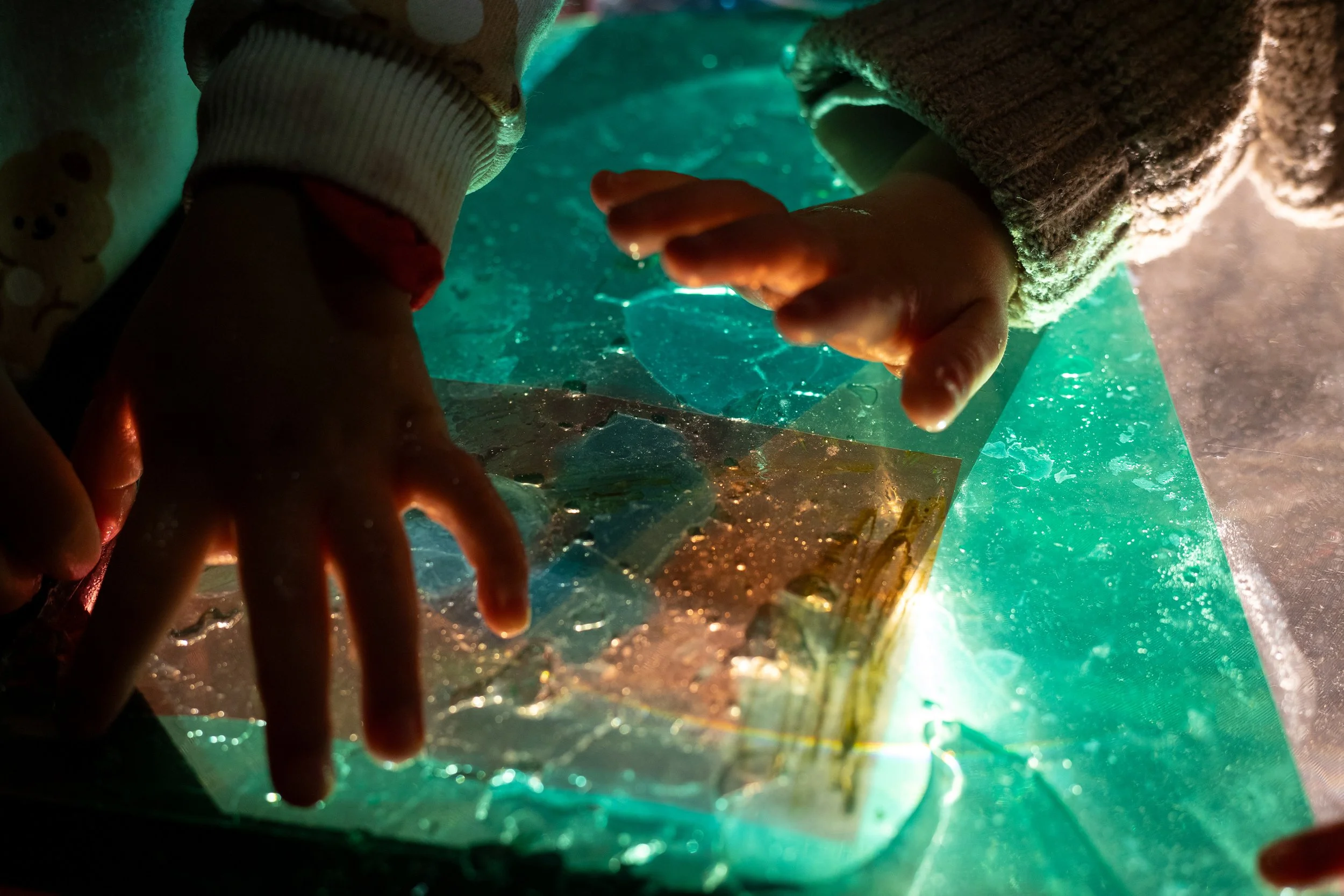 Mãos de duas crianças explorando uma superfície de vidro iluminada com luzes verdes e amarelas, observando pedaços de vidro transparente e pequenos objetos sobre ela.