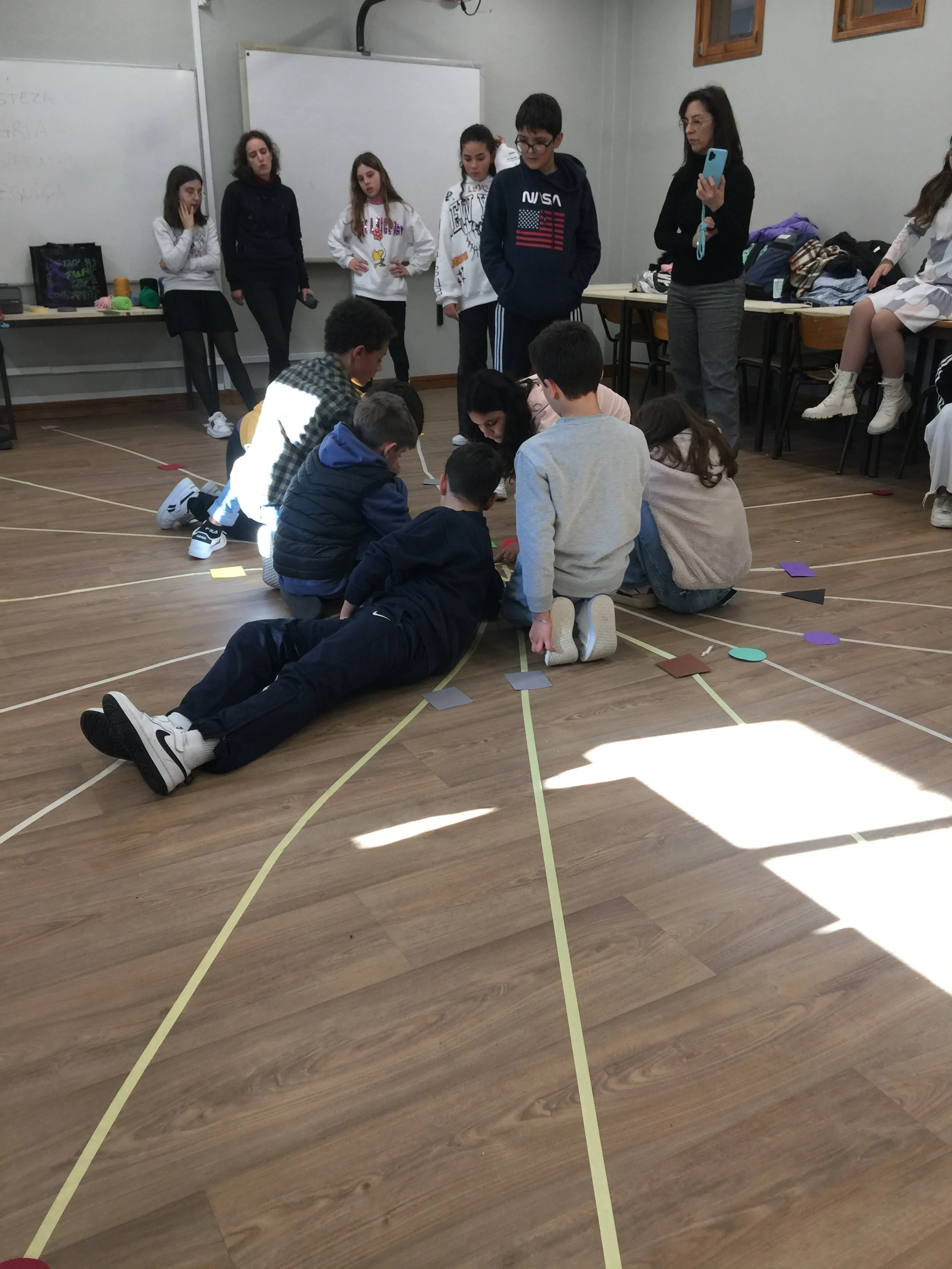 Grupo de crianças e adultos participando de uma atividade em uma sala de aula, com várias pessoas observando e uma delas gravando com o celular.