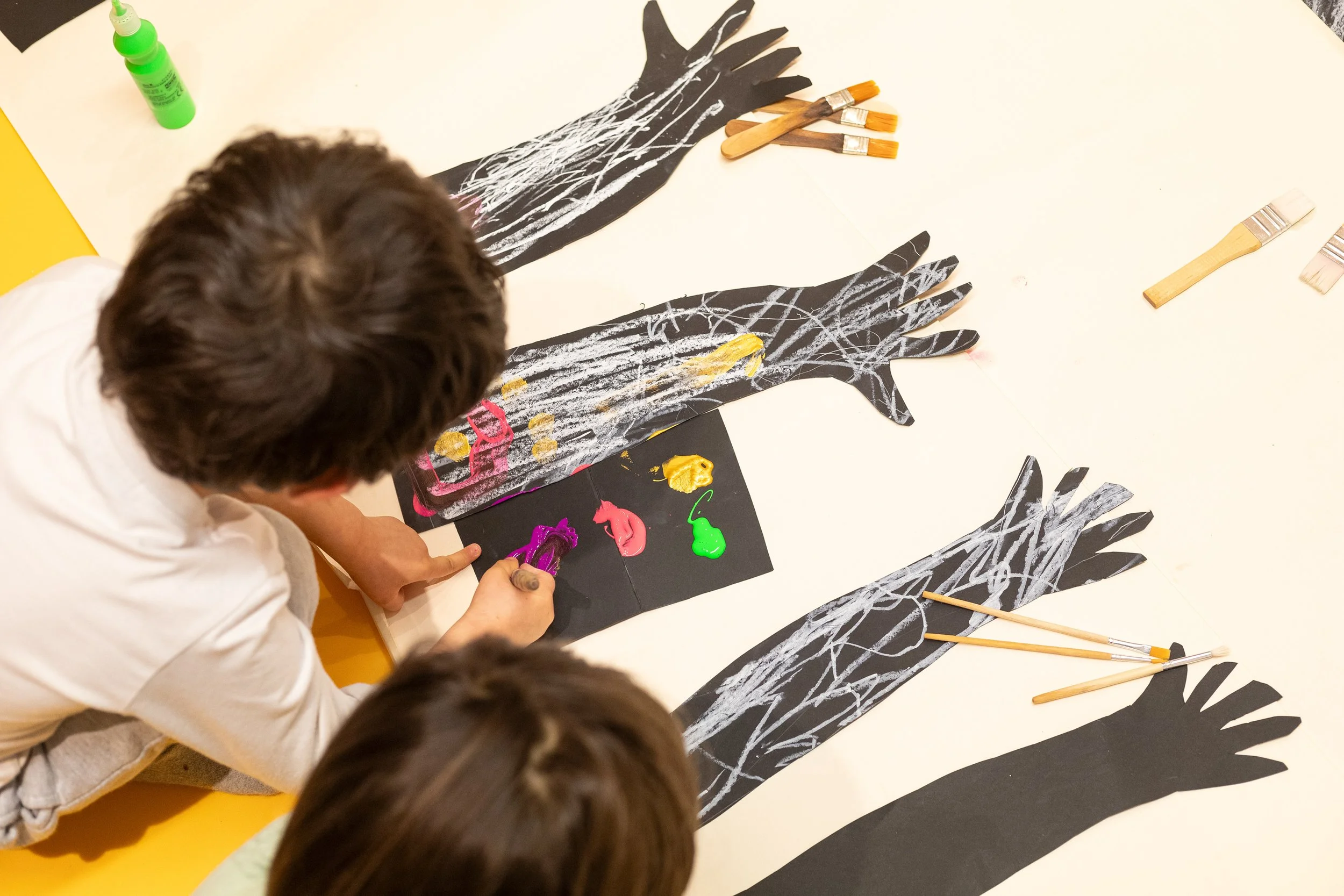 Crianças fazendo arte com folhas de papel preto recortadas em forma de mãos, usando tinta e pincéis para decorar.