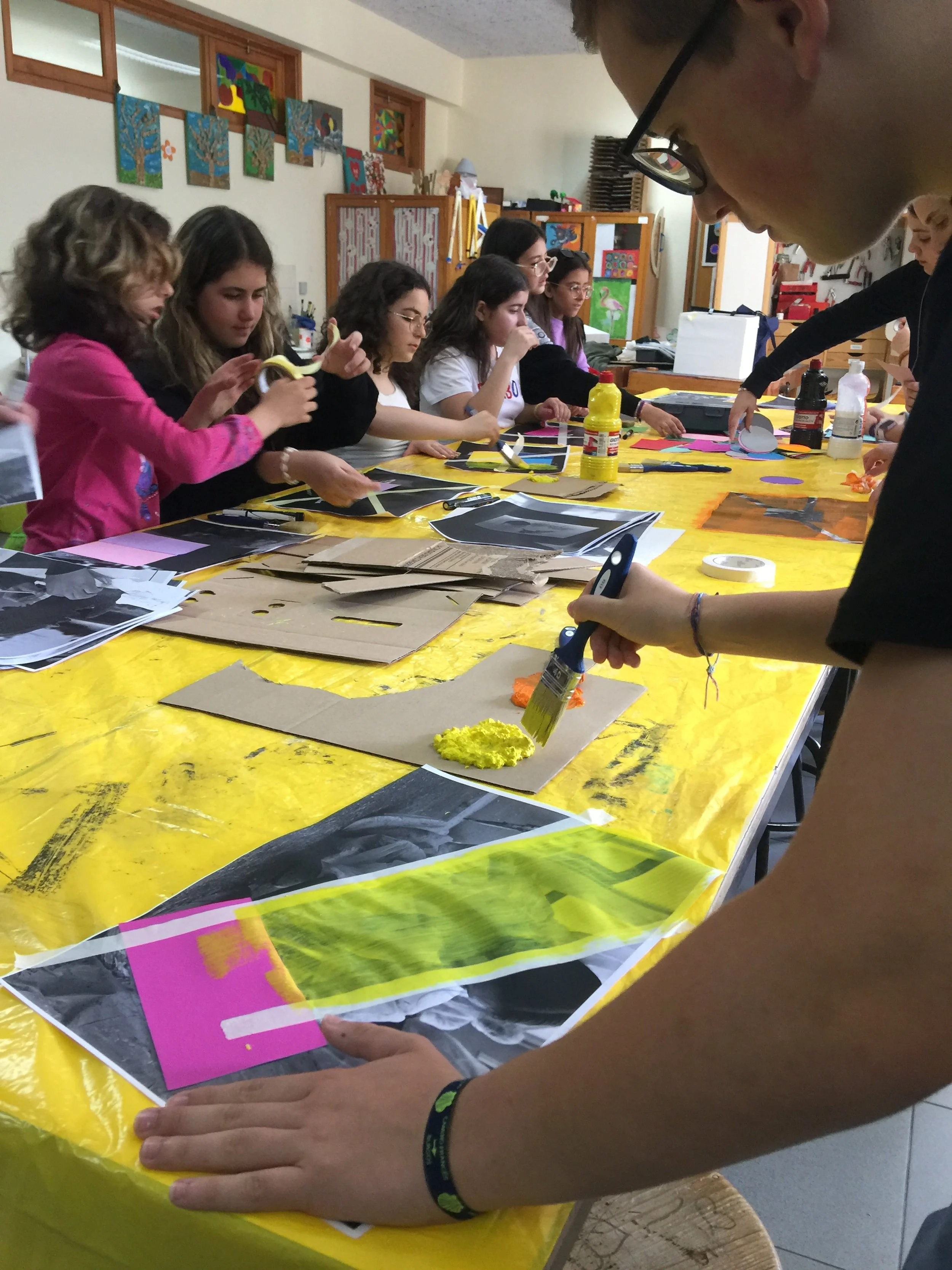 Grupo de pessoas em uma oficina de arte, pintando e utilizando papéis coloridos e fotografias em uma mesa amarela.