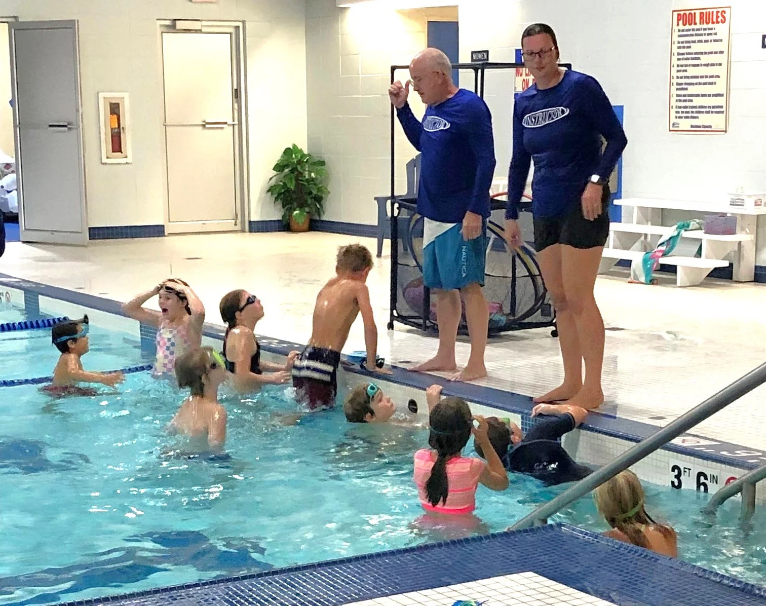A man and woman standing at the edge of the pool with children - swim lessons