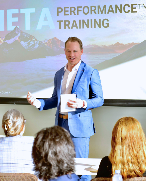 A man in a blue suit giving a presentation in front of a screen that reads 'IETA Performance Training.' He is smiling and gesturing to the audience, who are seated and listening attentively.