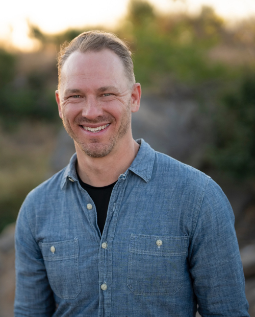 A man smiling outdoors wearing a blue button-up shirt, with a blurred natural background.