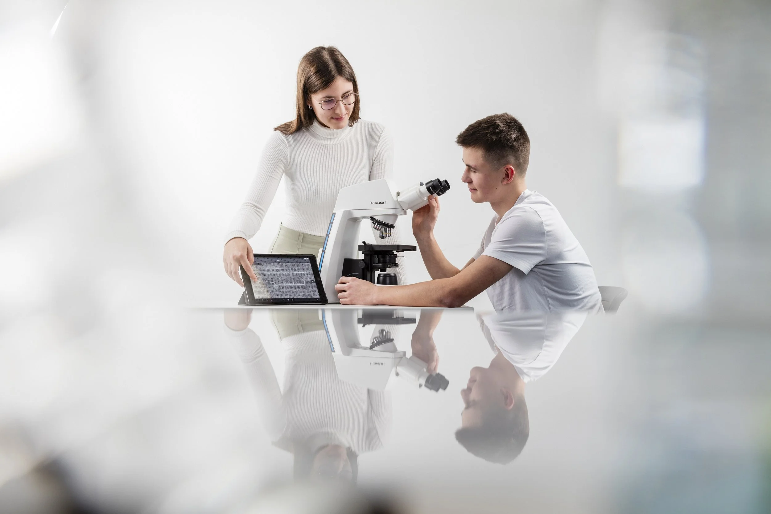 A woman and a young man in a laboratory, with the woman looking at a microscope and the young man seated, examining something through the microscope.