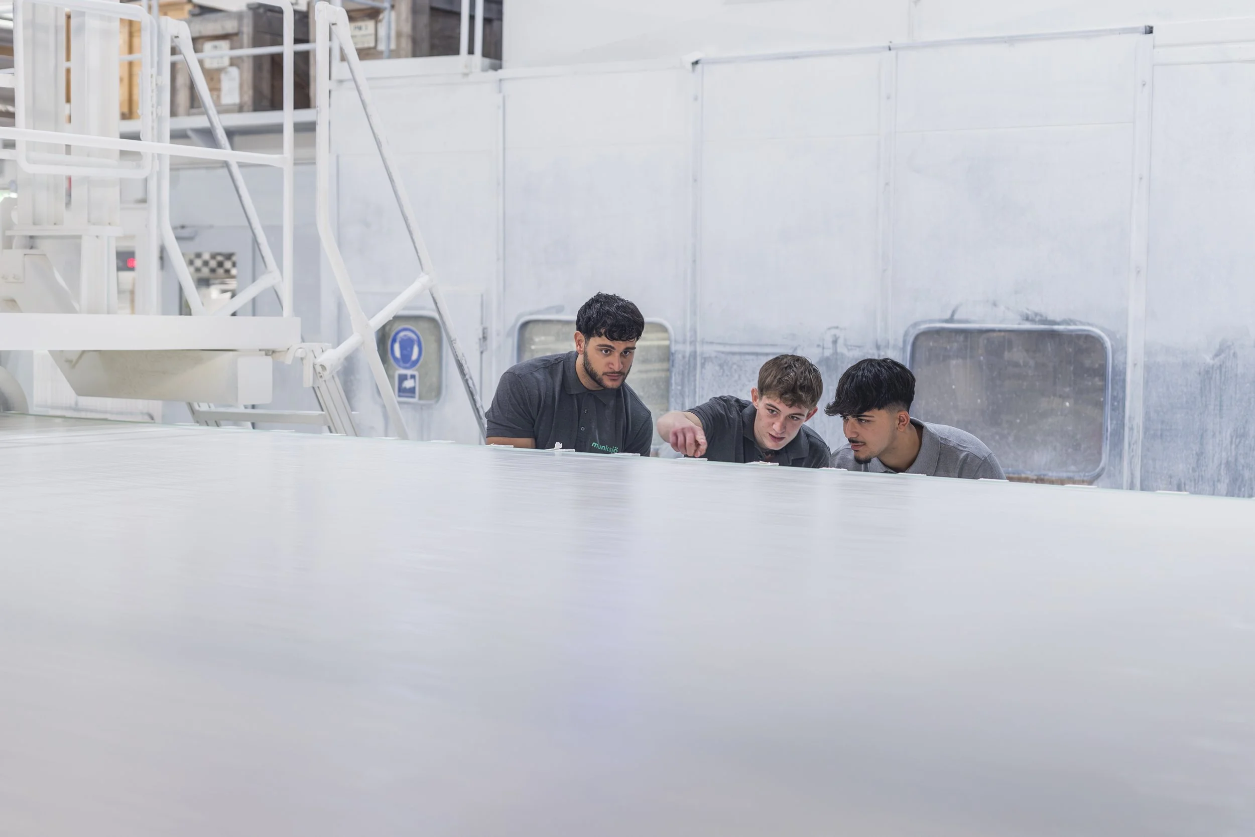 Three young men in work uniforms are inspecting a large, flat white surface in an industrial or manufacturing setting.