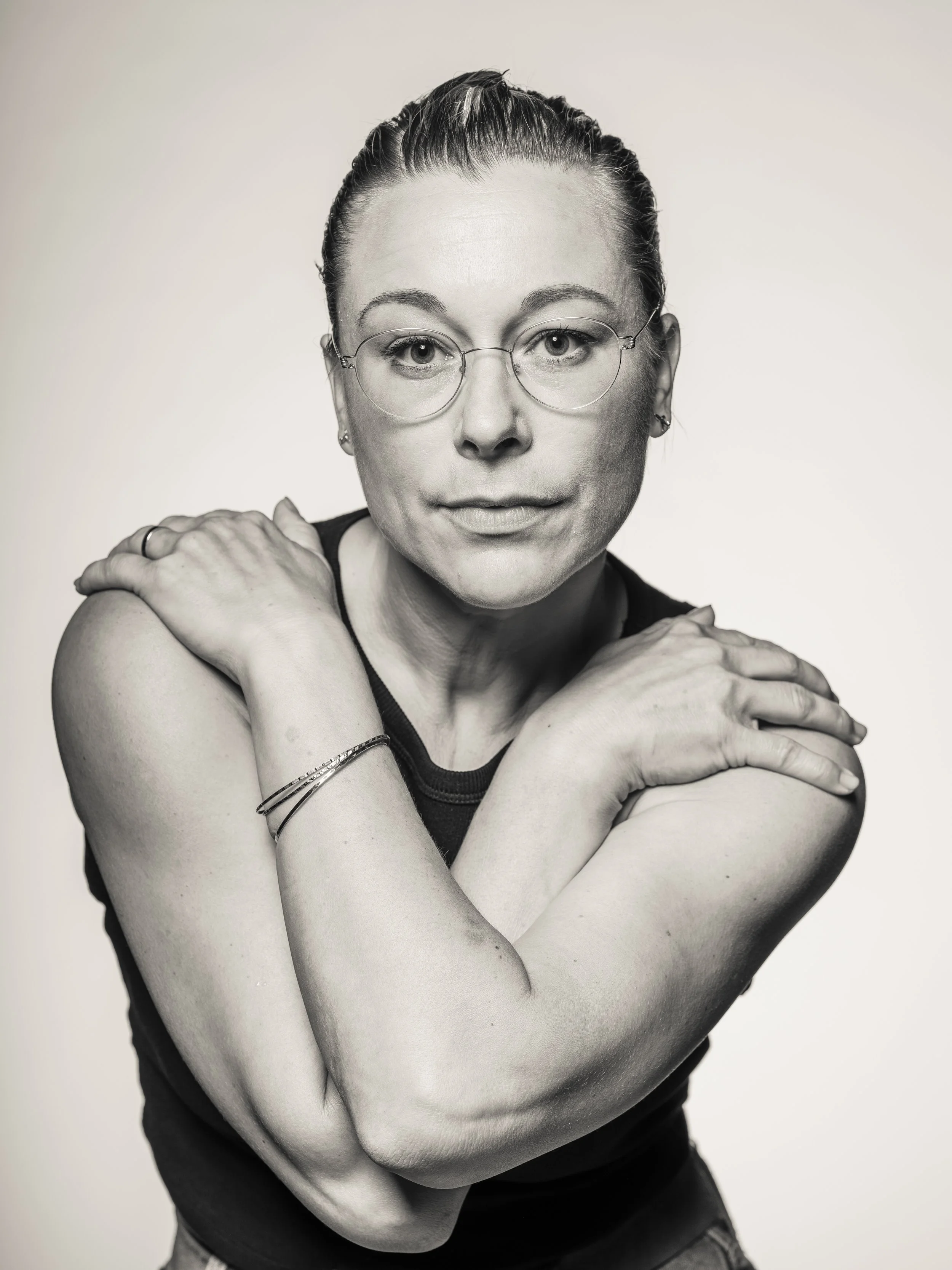 Black and white portrait of a woman with glasses, short hair, and jewelry, hugging herself with her arms crossed, against a plain background.