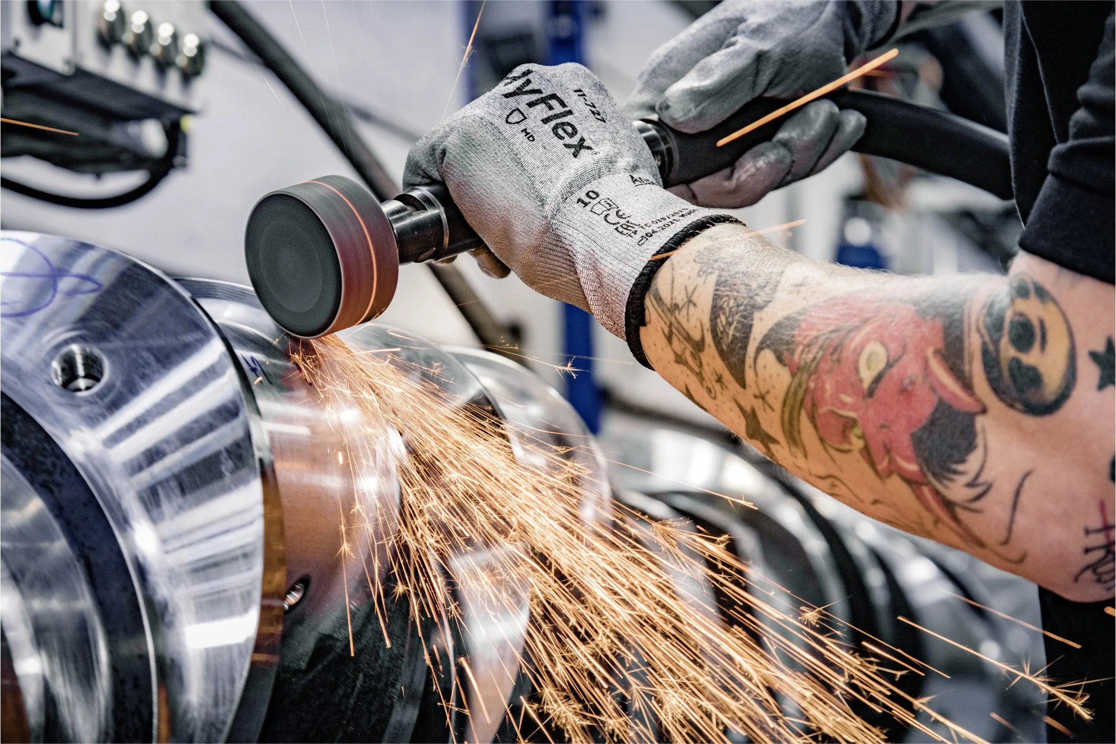A person with tattoos wearing gloves uses a grinding tool on a metallic part, creating sparks.