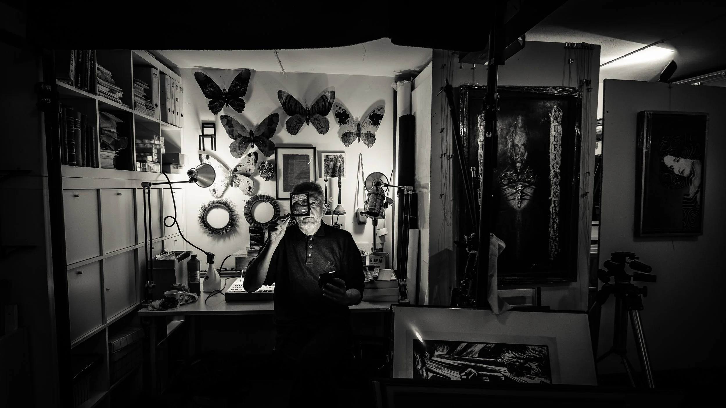 A man with white hair and a beard taking a selfie in a cluttered workshop or studio, with butterflies and framed pictures on the wall behind him, and various equipment and objects on the desk and shelves.