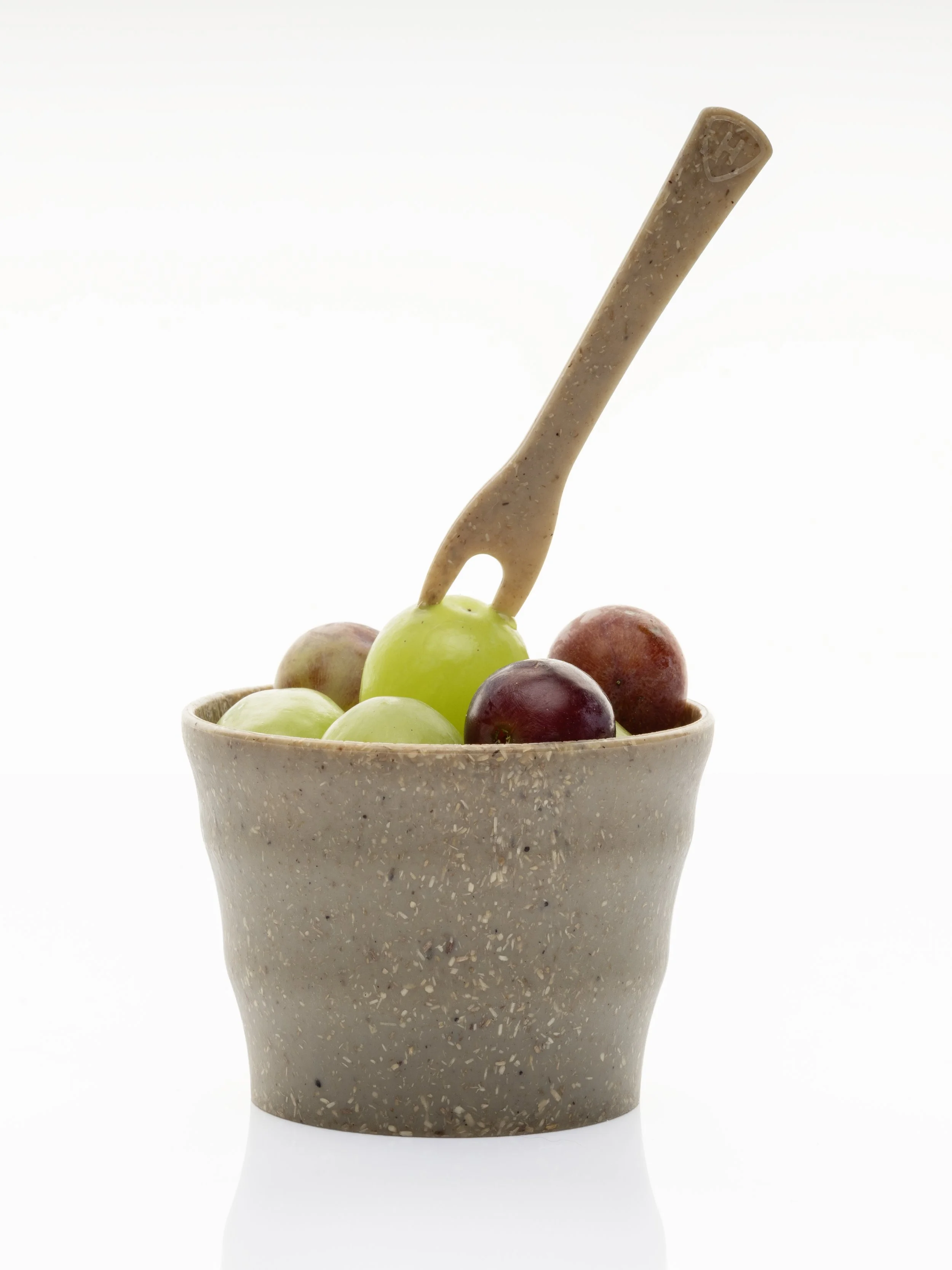A beige terrazzo vase filled with a variety of grapes, including green, red, and purple, with a wooden fork-like utensil inserted into the grapes, against a plain white background.