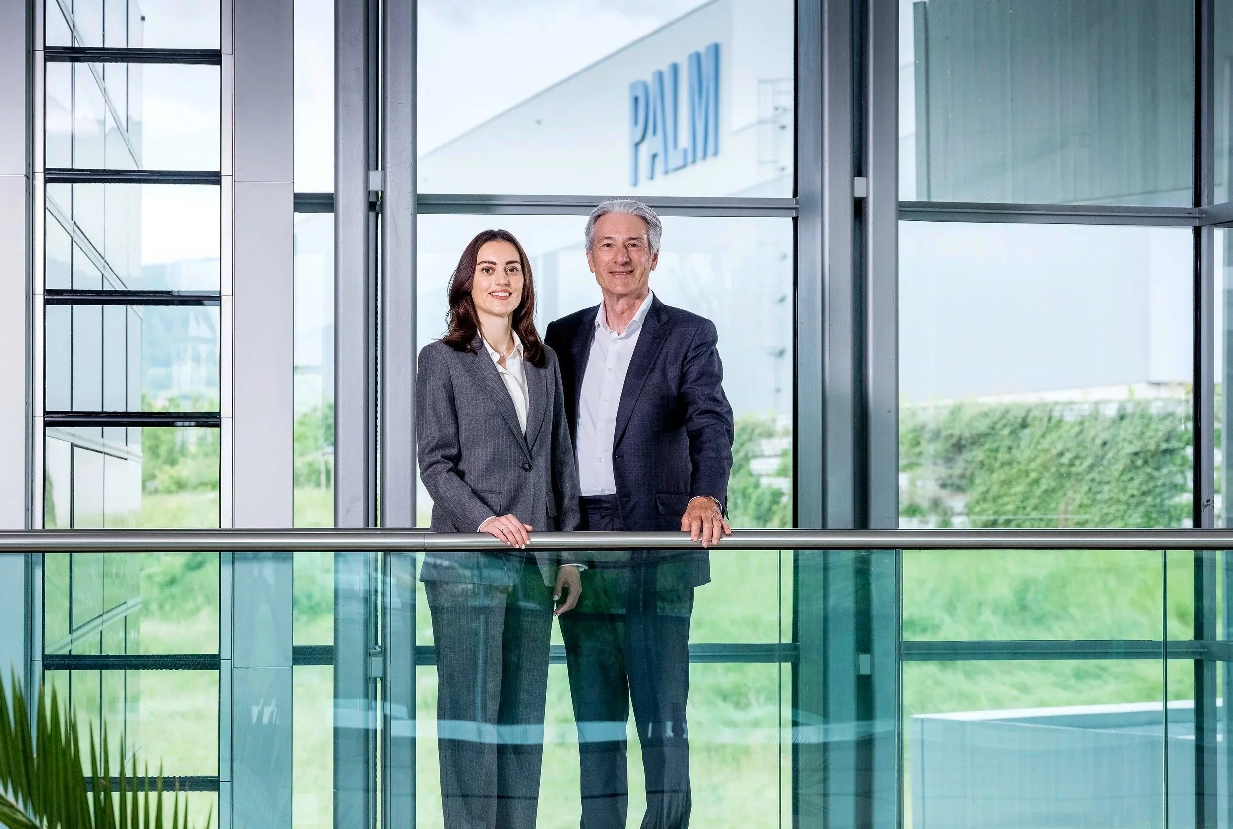 Two business professionals, a woman and a man, standing inside a modern office building near glass windows, with the signage 'PALM' visible outside