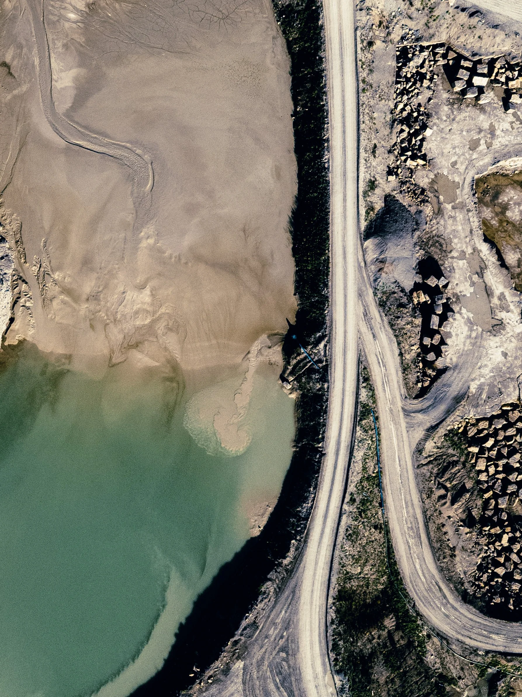 Aerial view of a winding road running alongside a large body of water with a muddy shoreline, rocky terrain, and sparse vegetation.